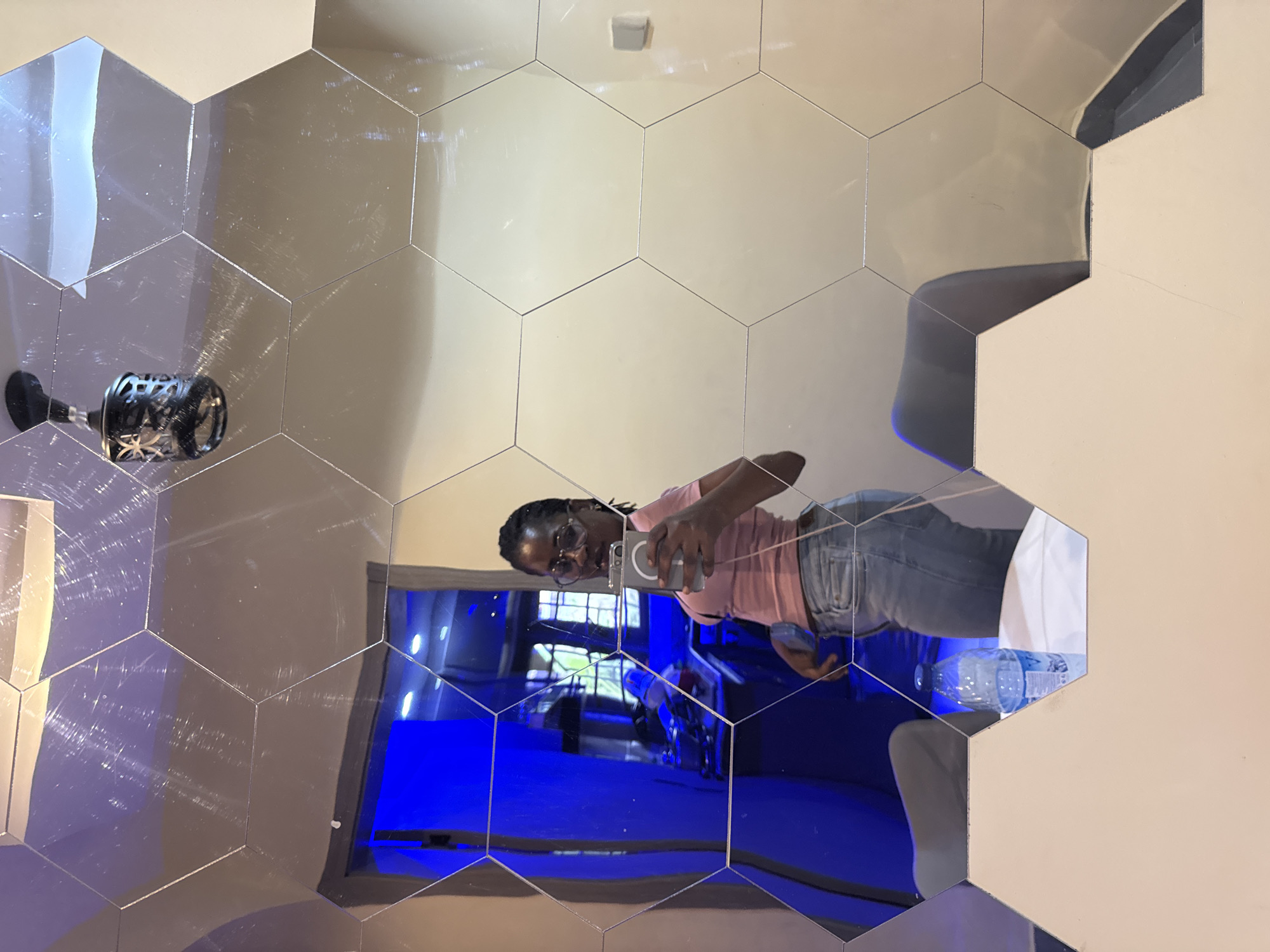 This is a mirror selfie taken in a room with hexagonal mirror tiles on the wall. The primary subject is a person reflected in the mirrors, appearing to be a young woman with braids and glasses, wearing a pink t-shirt and denim jeans. She is holding a smartphone and taking the photo. In the reflection, other objects are visible, including a black bottle with an intricate design, a water bottle, and a blue light source illuminating a section of the room. The overall setting appears to be indoors, with the hexagonal mirrors creating a fragmented and reflective environment. The lighting suggests it is daytime or the room is well-lit. The emotion conveyed is casual and self-aware, typical of a selfie. There is no visible text in the image.