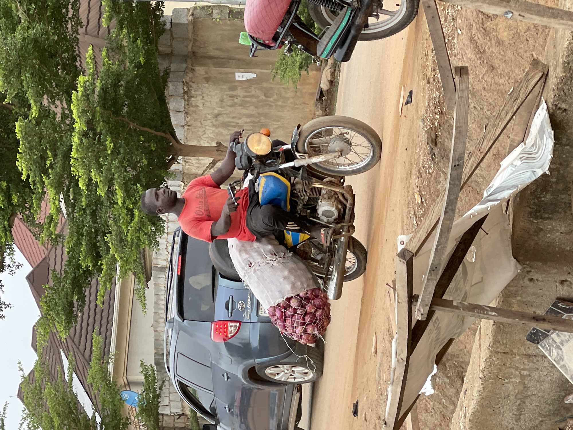 The image, oriented vertically, captures a street scene in Abuja, Nigeria, featuring a man on a motorcycle carrying a large load of produce.
The central subject is a man of dark complexion, wearing a bright red t-shirt with "Ohio State" visible on the shoulder and dark trousers. He is seated on a motorcycle, holding a small, silver object in his left hand, possibly a phone or a tool. The motorcycle is an older model, yellow and blue on the tank, with a prominent round headlight and visible engine components.
Attached to the rear of the motorcycle is a substantial, mesh-like sack brimming with red onions, extending from behind the rider over the back wheel. This suggests the man is transporting goods, likely for sale or delivery.
Behind the motorcycle, a dark gray Toyota RAV4 SUV is parked, with its "TOYOTA RAV4" emblem clearly visible on the rear hatch. Another motorcycle is partially visible further down the dirt road, which appears to be unpaved and dusty. To the left, a vibrant green tree and various bushes frame the scene, with a residential building featuring a brown tiled roof visible in the background. On the right side of the image, several crude wooden structures, resembling temporary stands, are erected, with some white fabric or tarp material attached to them.
The lighting indicates it is daytime, possibly overcast, as there are no distinct shadows. The man's expression is neutral, and the overall scene depicts a functional aspect of daily life and commerce.