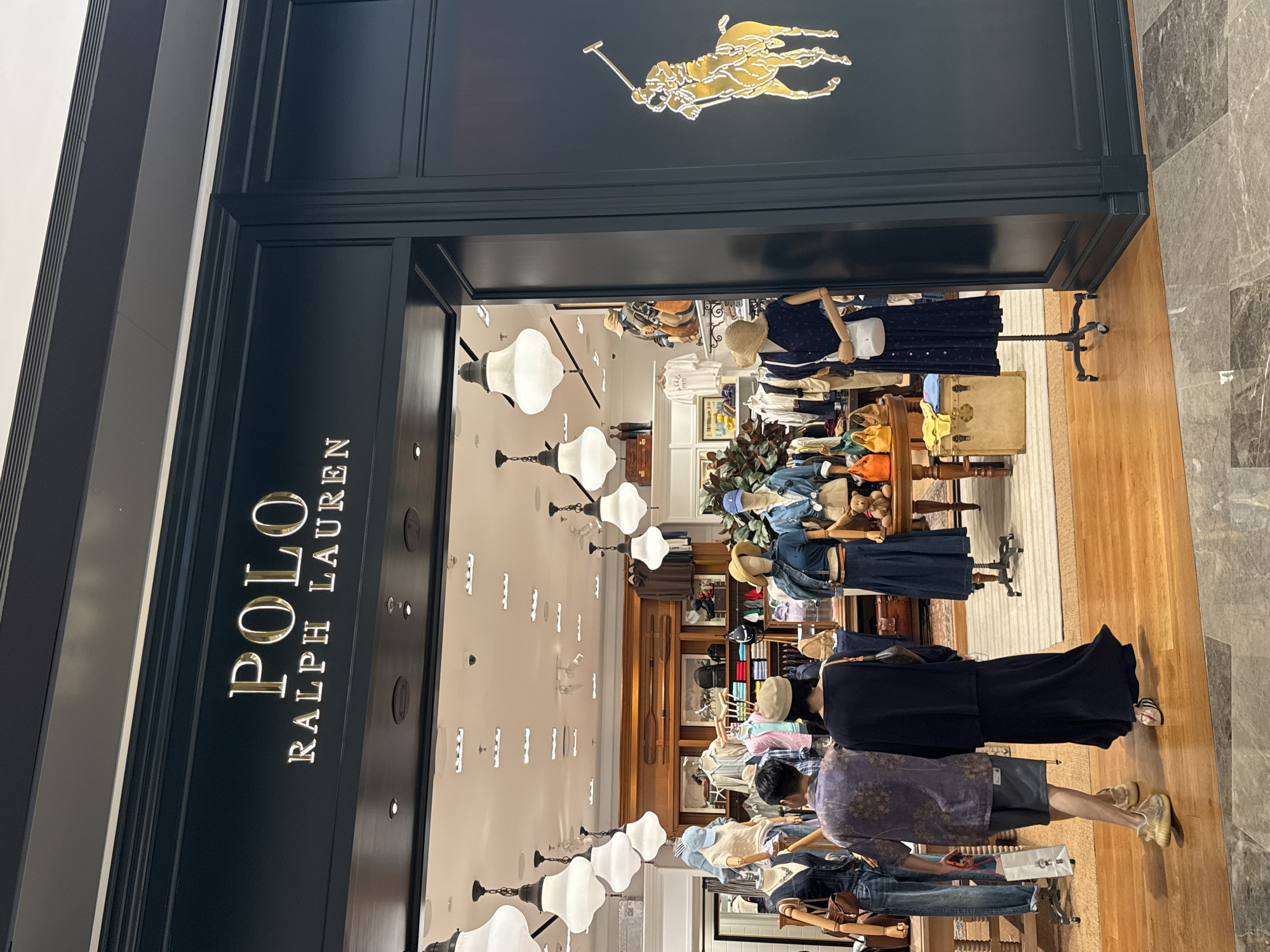 The image shows the exterior entrance of a Polo Ralph Lauren store in Liangjiang, China. The store facade is dark blue with gold lettering that reads "POLO RALPH LAUREN". A large gold Polo player logo is visible on the right side of the facade. Through the open entrance, a well-lit interior of the store can be seen, featuring clothing displays, mannequins dressed in various outfits, and wooden shelving. Two individuals, a male and a female, are walking away from the camera and into the store. The male is wearing a short-sleeved patterned shirt and shorts, carrying a white shopping bag. The female is wearing a dark long dress and carrying a handbag. The floor in front of the store is a mix of wood and marble tiling. Several large, decorative pendant lights hang from the ceiling inside the store. The overall scene depicts a retail environment with customers entering the establishment.