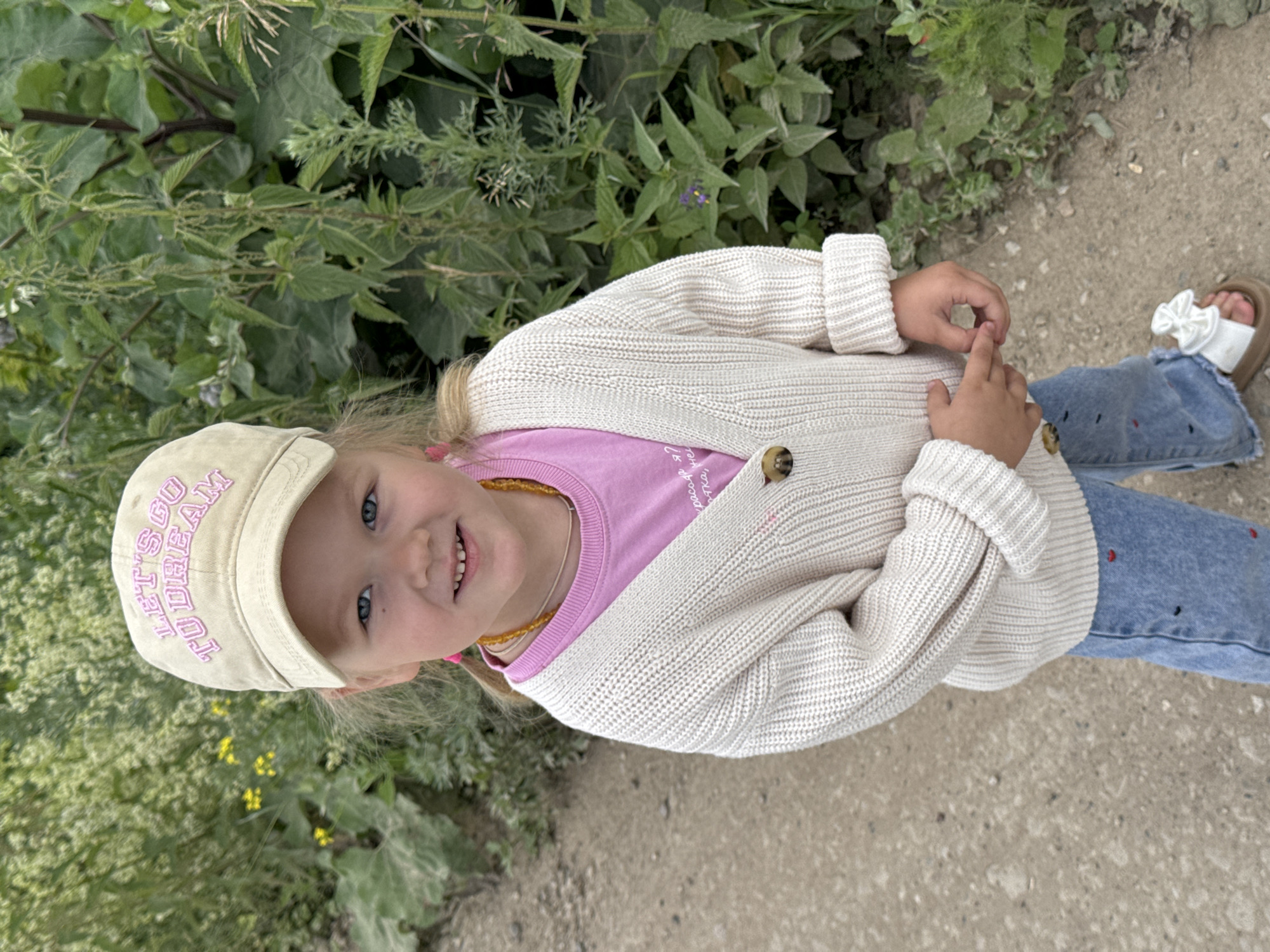 A young girl with blonde hair, blue eyes, and a bright smile is pictured outdoors. She is wearing a beige baseball cap with the words "LET'S GO TO DREAM" embroidered in purple letters. She has on a pink t-shirt and a cream-colored knitted cardigan with two brown buttons. Around her neck, she wears a yellow beaded necklace. Her lower half is covered by light blue denim jeans adorned with small red and black embroidered shapes, and a pair of brown sandals with white bows.
The girl is sitting on the ground, which appears to be a dirt path. Behind her and to her left, lush green foliage and plants with small yellow flowers are visible, suggesting a natural, possibly rural or park-like setting. The lighting indicates it is daytime, with soft, diffused light. The overall mood is cheerful and relaxed, with the girl looking directly at the camera with a happy expression.
The provided context specifies the location as "Горный, Russia." There is no specific information about the time of day or weather beyond the visual cues of daylight.