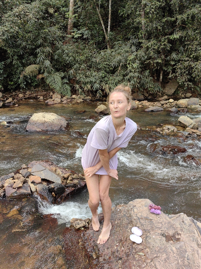 A woman stands on a rock in a river, wearing a light purple t-shirt. The river flows around her, surrounded by rocks and greenery. She has her hair pulled back in a bun and is looking off to the side. Her bare feet are on the rock next to a pair of white sandals and colorful flip-flops. The background is a dense forest. The light is diffused.