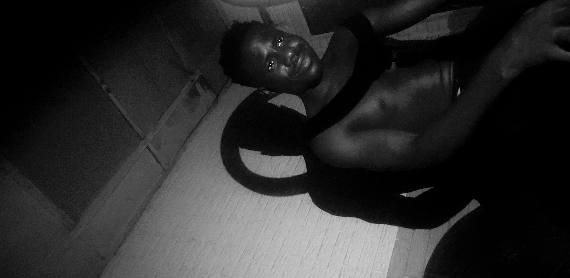 A black and white photograph, likely taken indoors in Narayi, Nigeria, depicts a man lying on a tiled surface. The man has dark skin and short hair. He appears shirtless and is looking directly at the camera with a slight smile. A dark, unidentified object is draped across his chest, creating shadows that obscure portions of his body. The background includes a partially visible window with a wooden frame. The overall lighting in the photograph is low, with strong contrasts between light and shadow.