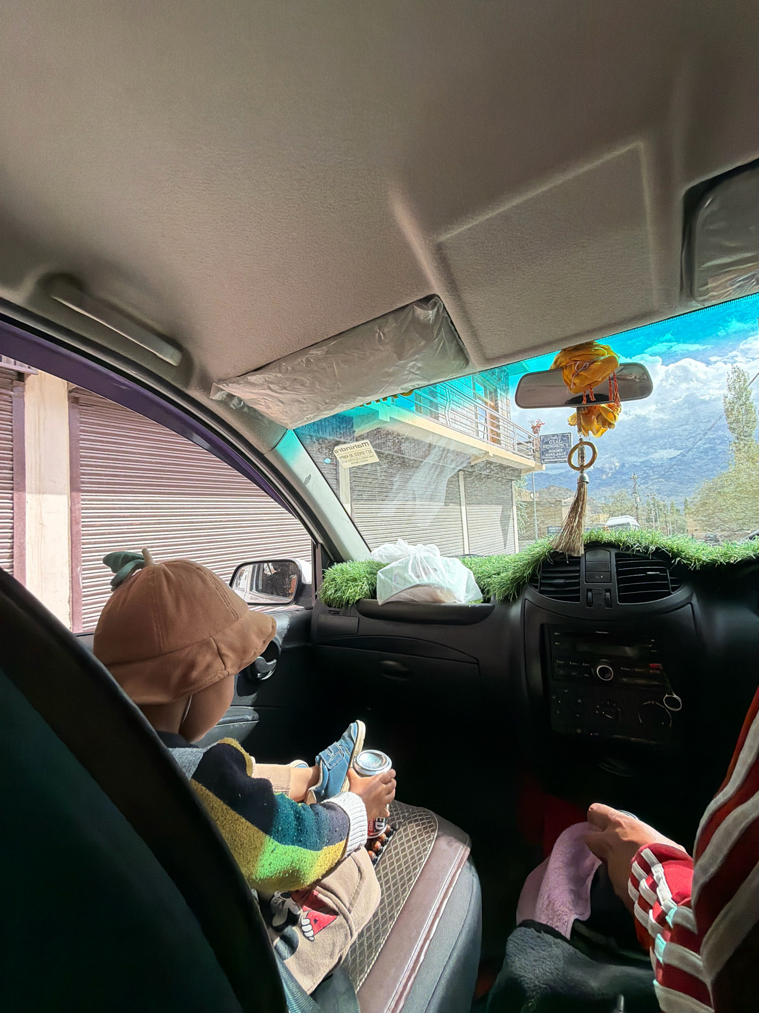 A passenger's-side view from inside a vehicle in Leh, India shows a child wearing a brown hat and a colorful striped sweater, sitting with their feet up, holding a can. The dashboard is decorated with artificial grass and a yellow tassel ornament hangs from the rearview mirror. Outside the car, buildings with roll-up doors and a mountain landscape under a cloudy sky are visible. The scene suggests a travel or commute within the city.