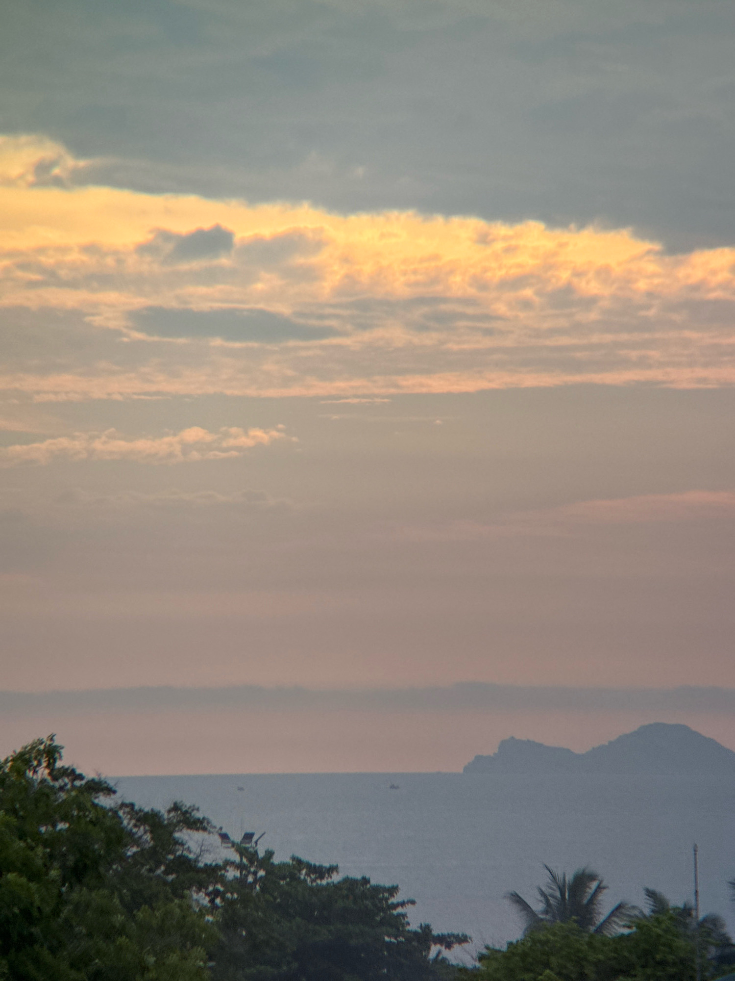 The image depicts a serene coastal landscape, likely taken from a vantage point overlooking the ocean in Nha Trang, Vietnam. In the foreground, lush green foliage, including trees and palm fronds, fills the bottom portion of the frame, suggesting a tropical environment.
Beyond the trees, a body of water stretches to the horizon, appearing calm and slightly hazy. A few distant boats are visible on the water, their presence indicating some maritime activity. Silhouetted against the sky in the distance is a chain of islands or a large headland, their shapes softened by atmospheric perspective.
The sky is a dominant feature of the image, displaying a gradient of colors characteristic of either sunrise or sunset. The upper portion of the sky is a muted blue-gray, transitioning into bands of soft orange and pale pink as it moves lower. wispy clouds are scattered throughout, catching the light and adding texture to the sky. The overall impression is one of tranquility and natural beauty.
