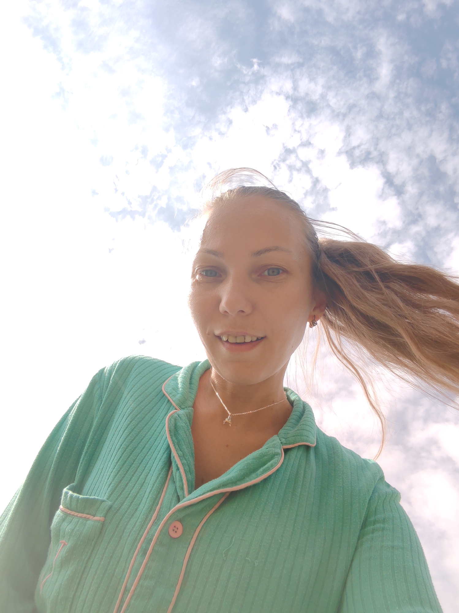 A low-angle, waist-up selfie shows a woman with blonde hair in pigtails, looking up at the camera with a smile. She is wearing a light teal pajama set with pink trim and buttons. A silver necklace with a star pendant is visible around her neck. Her hair is being blown by the wind, creating a dynamic effect.
The background is a bright, cloudy sky, suggesting it is daytime. The clouds are white and fluffy, with some darker grey patches, indicating a mix of sun and clouds. The lighting is strong, with the sun appearing to be behind and to the left of the subject, causing some lens flare and overexposure in the upper left corner of the image.
The setting appears to be outdoors, possibly in a backyard or an open area, given the exposure to the sky and wind. The overall mood is casual and cheerful, conveyed by the woman's smile and the bright, open sky. There is no visible text in the image.