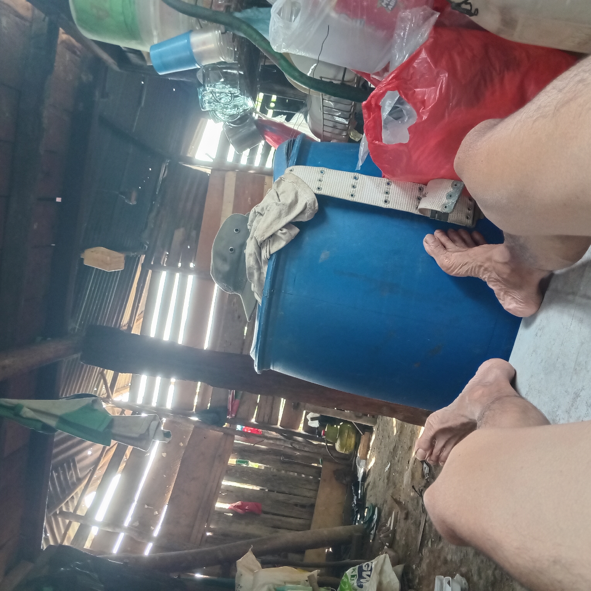 The image shows a close-up view of a person's legs and feet resting on a white surface, possibly a mat or bedsheet. The background is a cluttered indoor space with a rustic, makeshift appearance. Sunlight streams in through gaps in the corrugated metal roof and wooden walls, creating bright rays. Various objects are visible, including a large blue barrel with a hat and clothing draped over it, a green gas tank, a red plastic bag, and other containers and items scattered on the dirt floor. Wooden beams and planks form the structure of the shelter. The overall impression is of an informal, possibly rural or temporary living environment.