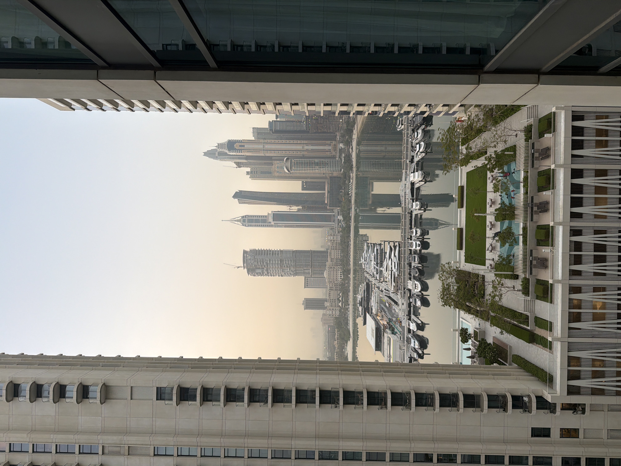 The image captures a high-angle view of the Dubai Marina in the United Arab Emirates. On the left, a tall, beige apartment building with numerous balconies and windows dominates the foreground. On the right, another modern building partially frames the scene, revealing its facade with reflective glass windows and balconies.
Below these buildings, a large marina filled with numerous yachts and smaller boats is visible. The boats are neatly docked and reflect on the calm water. Beyond the marina, a lush green rooftop terrace with lounge chairs, umbrellas, and trees is situated on top of a lower building. Further in the distance, the iconic skyscrapers of the Dubai skyline rise against a hazy sky, their reflections shimmering on the water. The overall atmosphere is one of urban luxury and a bustling waterfront.