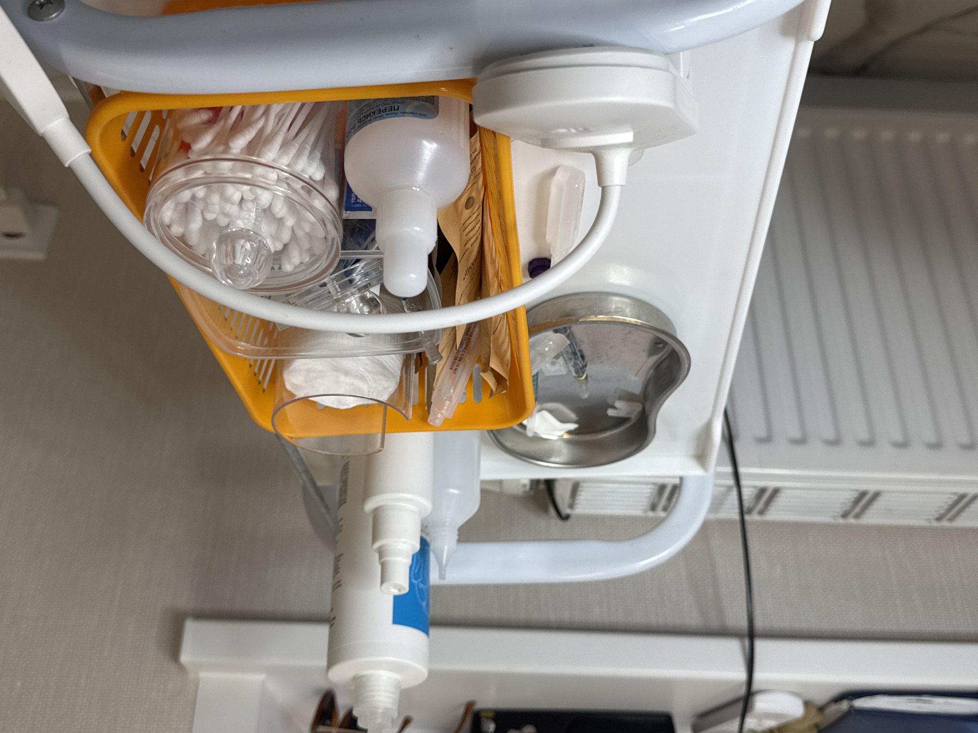 This is a forensic, matter-of-fact description of an image depicting a medical trolley in what appears to be a medical or cosmetic treatment room. The trolley is white with a grey handle. On the trolley, various medical supplies are organized in an orange plastic basket. These supplies include several bottles of liquids, syringes, cotton swabs in a clear container, and a metal tray containing what appears to be a syringe and gauze. There is also a white device with a flexible white tube attached to it. The background shows a radiator, a white shelf with some electronic devices and papers, and a beige textured wall. The lighting suggests an indoor environment. The location in Perm, Russia is not explicitly indicated by any visible markers in the image.