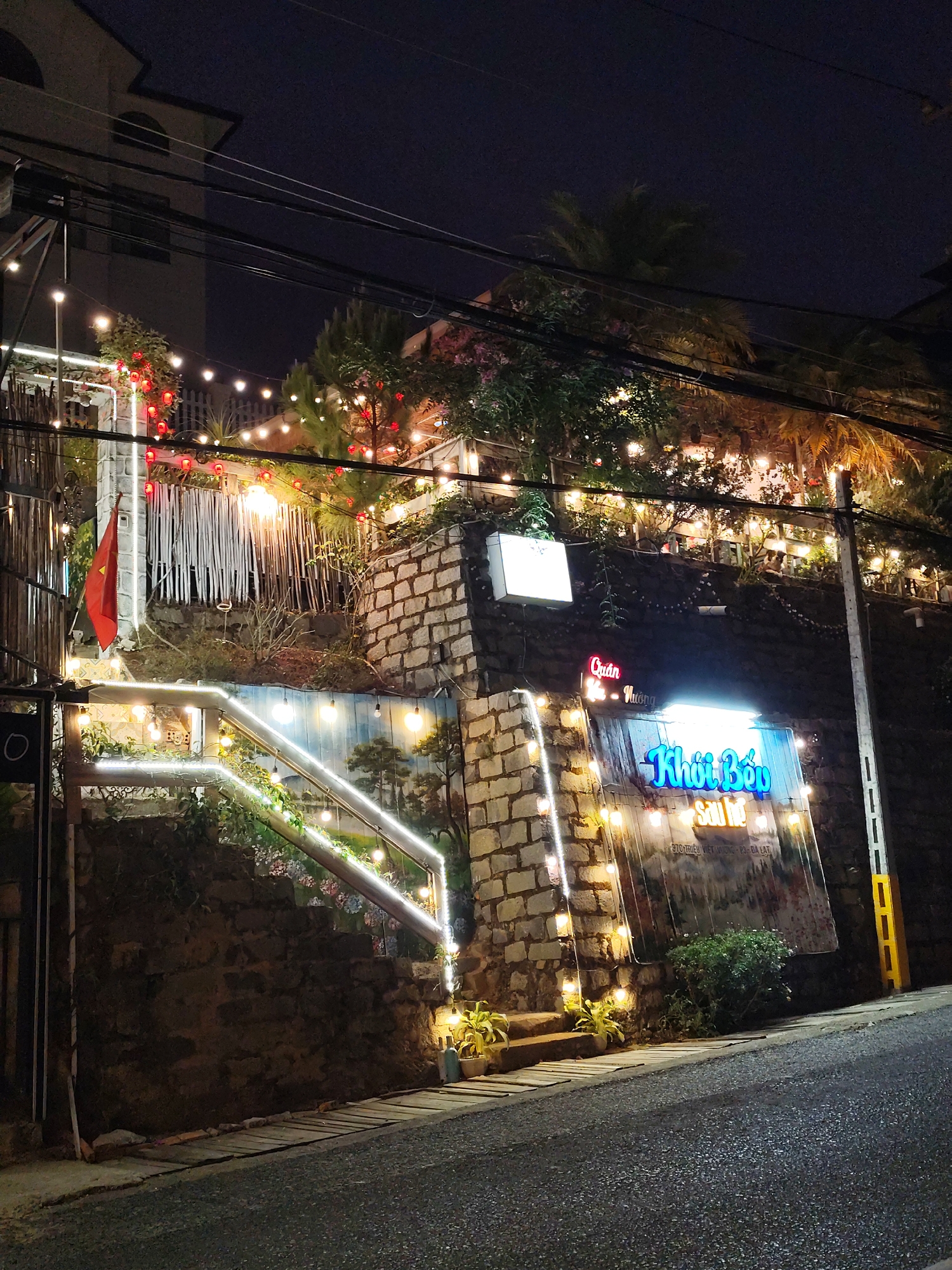 A nighttime street scene in Da Lat, Vietnam, shows a hillside business, likely a restaurant or bar, decorated with strings of lights. The building's facade is constructed of stone and partially obscured by vegetation. A brightly lit sign reading "Khởi Bếp Sau Hồ" is visible, along with a smaller sign above that reads "Quán - Nướng." A Vietnamese flag is prominently displayed. The street is paved and slightly inclined. Utility poles and wires are visible overhead. The overall atmosphere is festive and inviting.