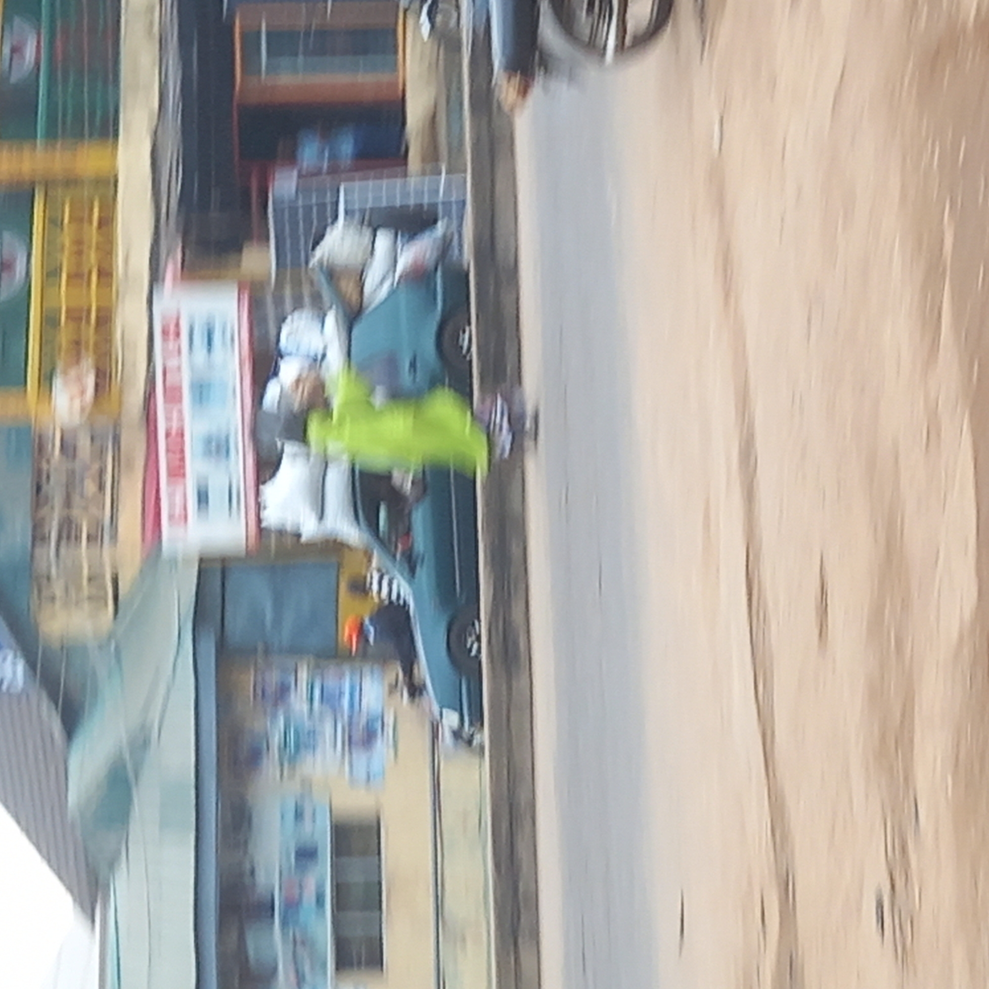 The image shows a street scene in Dadi Riba, Nigeria. A person wearing a bright green outfit is walking along the sidewalk. A turquoise car is parked nearby. Buildings with storefronts are visible in the background, along with signage in local languages. A motorcycle is partially visible at the top of the frame. The road surface is light brown.