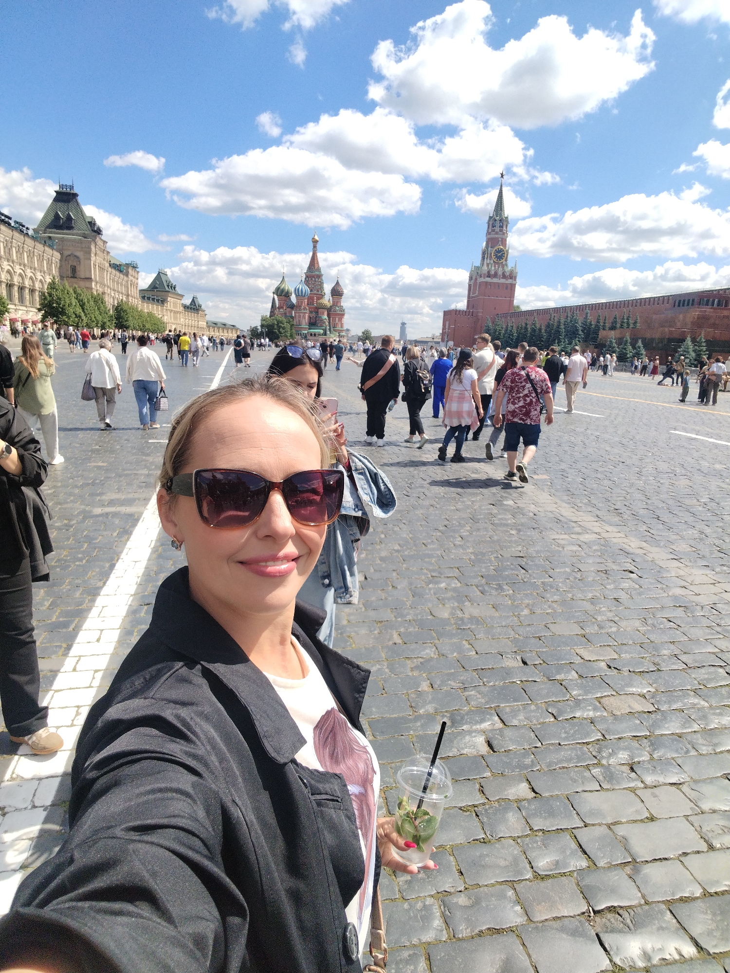 A woman is taking a selfie in Red Square, Moscow, Russia, on a sunny day with a few clouds in the sky. She is smiling and wearing sunglasses, a black jacket over a white t-shirt with a graphic print, and holding a clear cup with a drink and mint leaves. In the background, there are many tourists walking on the cobblestone square. Prominent landmarks visible include the State Historical Museum on the left, St. Basil's Cathedral in the middle, and the Spasskaya Tower of the Moscow Kremlin on the right.