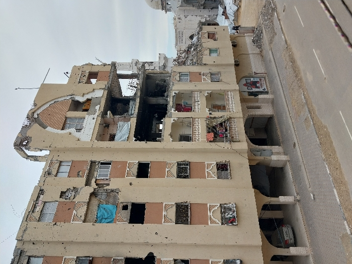 This image displays a multi-story building in Khan Yunis, Palestinian Territory, exhibiting extensive structural damage under an overcast sky. The light-beige structure, featuring brown decorative elements and arched ground-floor entries, shows significant collapse on its upper right side, with the facade and interior floors largely missing and rebar exposed. Numerous projectile impacts and holes are visible across the entire visible facade. Windows are broken or absent. Blue fabric hangs from a damaged window opening on the left side, and various items of clothing are draped over railings on a third-floor balcony. A white sedan is parked underneath the building's ground-floor arches on the left. A red and white commercial sign, partially obscured, reading "O&M" and "TECHNET" is visible on the ground floor. Piles of debris are present on the ground adjacent to the building on the right, where two small, dark figures are discernible. An asphalt road with white painted lines and a paved sidewalk are in the foreground. In the background, to the right, a white-domed structure, consistent with a mosque, and other heavily damaged buildings are visible amidst further rubble.