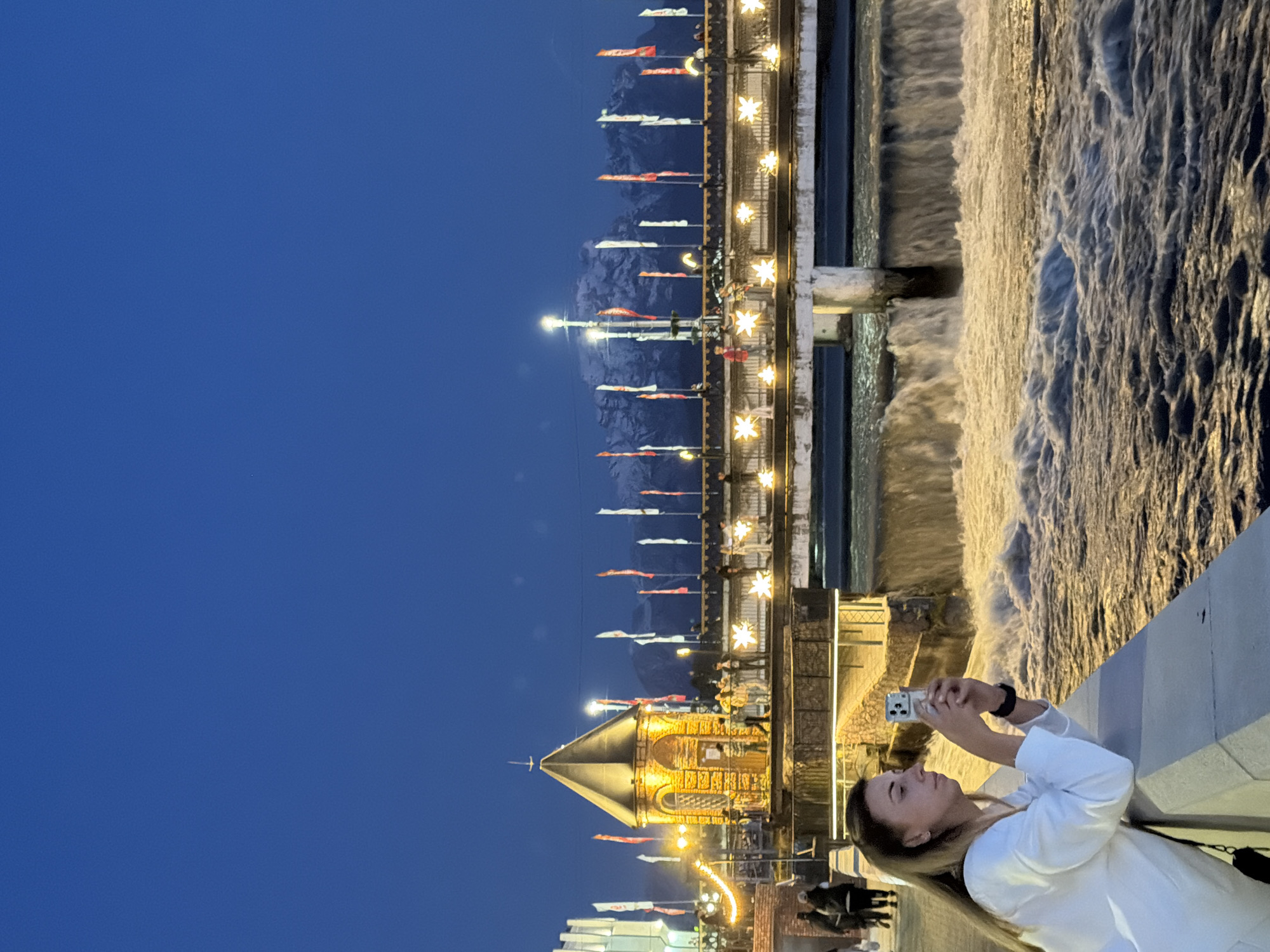 A long shot captured outdoors at dusk shows a woman taking a photo of the Chermenskiy waterfall in Chermen, Russia. A woman in a white shirt is standing on a bridge, holding a smartphone up to take a photo, as a wide waterfall rushes over the structure in the background. Above the waterfall is a walkway with a line of colorful pennants. Beyond that, dark mountains rise in the distance. To the left of the waterfall, a short stone structure with a gold spire and lit windows is also visible. Below the stone structure, a modern white building and other buildings with lights on are present. The sky is a deep blue, suggesting early evening. There are also other people present further away on the walkway and near the woman.