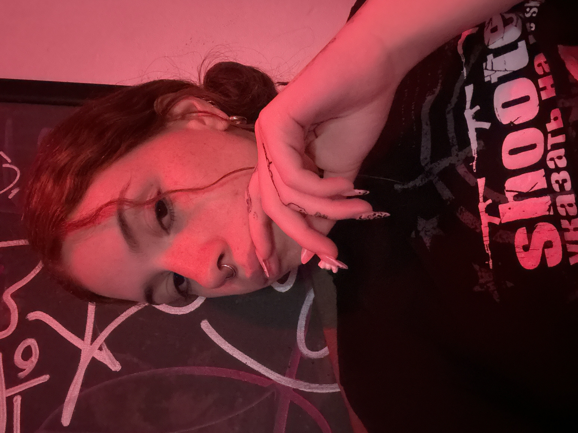 A person lies on a surface patterned with white and purple markings. They have light skin, freckles, and brown hair pulled back into a bun. They wear a black t-shirt with white and red text. The person's right hand is near their face, with patterned nails. The image is dominated by a red light. The location is unknown and cannot be determined from the image.