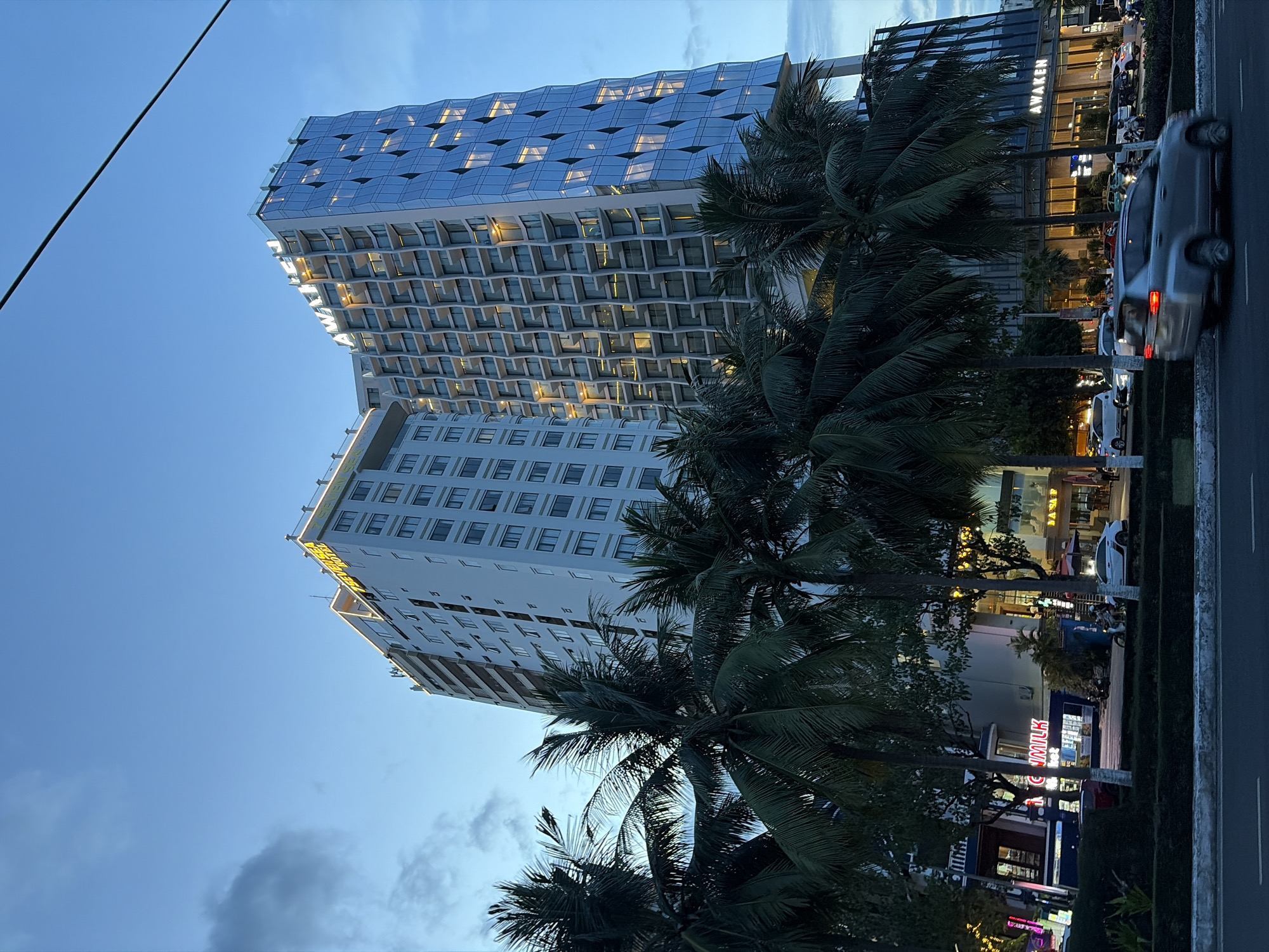 In the city of Đà Nẵng, Vietnam, an evening scene unfolds with two prominent modern buildings dominating the skyline. The building on the left is a tall, rectangular structure with a light-colored facade, adorned with the glowing yellow text "SEAVIEW HOTEL" along its upper edge. Adjacent to it, the building on the right features a more contemporary design with a facade composed of blue glass panels arranged in a diamond pattern, reflecting the twilight sky. Both structures are illuminated, with warm lights visible from within the windows. In the foreground, lush palm trees with dark green fronds create a tropical atmosphere, partially obscuring the lower levels of the buildings. A street runs in front of the buildings, with a silver car in motion in the foreground, its taillights illuminated. Several other vehicles are visible on the street and parked along the side. The sky above is a dusky blue with scattered clouds. The overall impression is one of a vibrant urban setting with a blend of modern architecture and tropical vegetation.