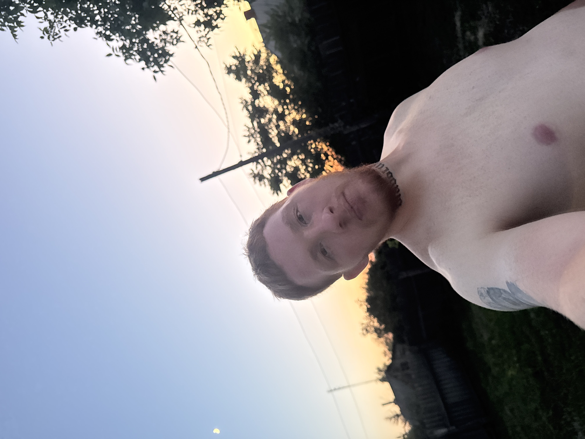 A shirtless man with short, reddish-brown hair and a beard is taking a selfie outdoors. He has a tattoo on his neck. The background shows a fence, trees, and houses against a sunset sky with a crescent moon visible. The man is positioned in the foreground, with his upper body filling most of the frame. The lighting suggests it is dusk. The location is described as Unknown, Unknown.