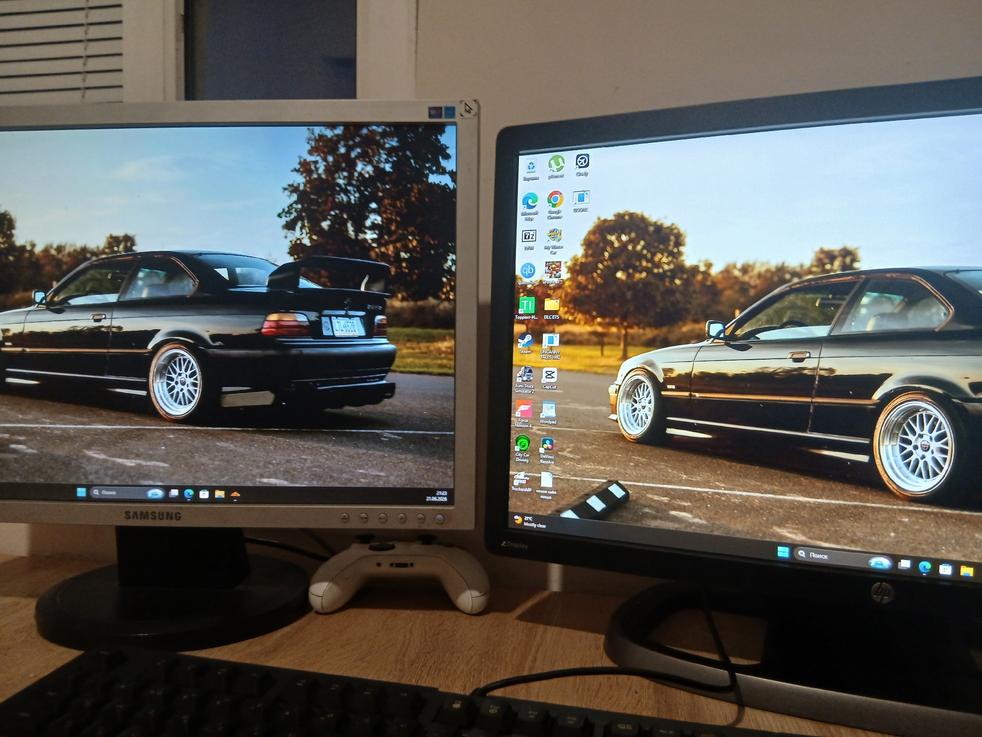 The image shows a desk setup with two computer monitors displaying wallpapers of a black BMW M3 E36. The left monitor, a Samsung brand, shows the rear of the car, while the right monitor, an HP brand, shows the side profile of the car. Both monitors are displaying a desktop environment with various icons and a taskbar. A white video game controller is placed in front of the left monitor. A black keyboard is visible in the foreground. The environment appears to be indoors, likely a home office or gaming room, with a wooden desk surface. The wallpaper suggests a preference for performance cars. There are no people or animals visible in the image. The scene does not provide any specific visual cues that would allow for identification of the location in Dnipro, Ukraine.