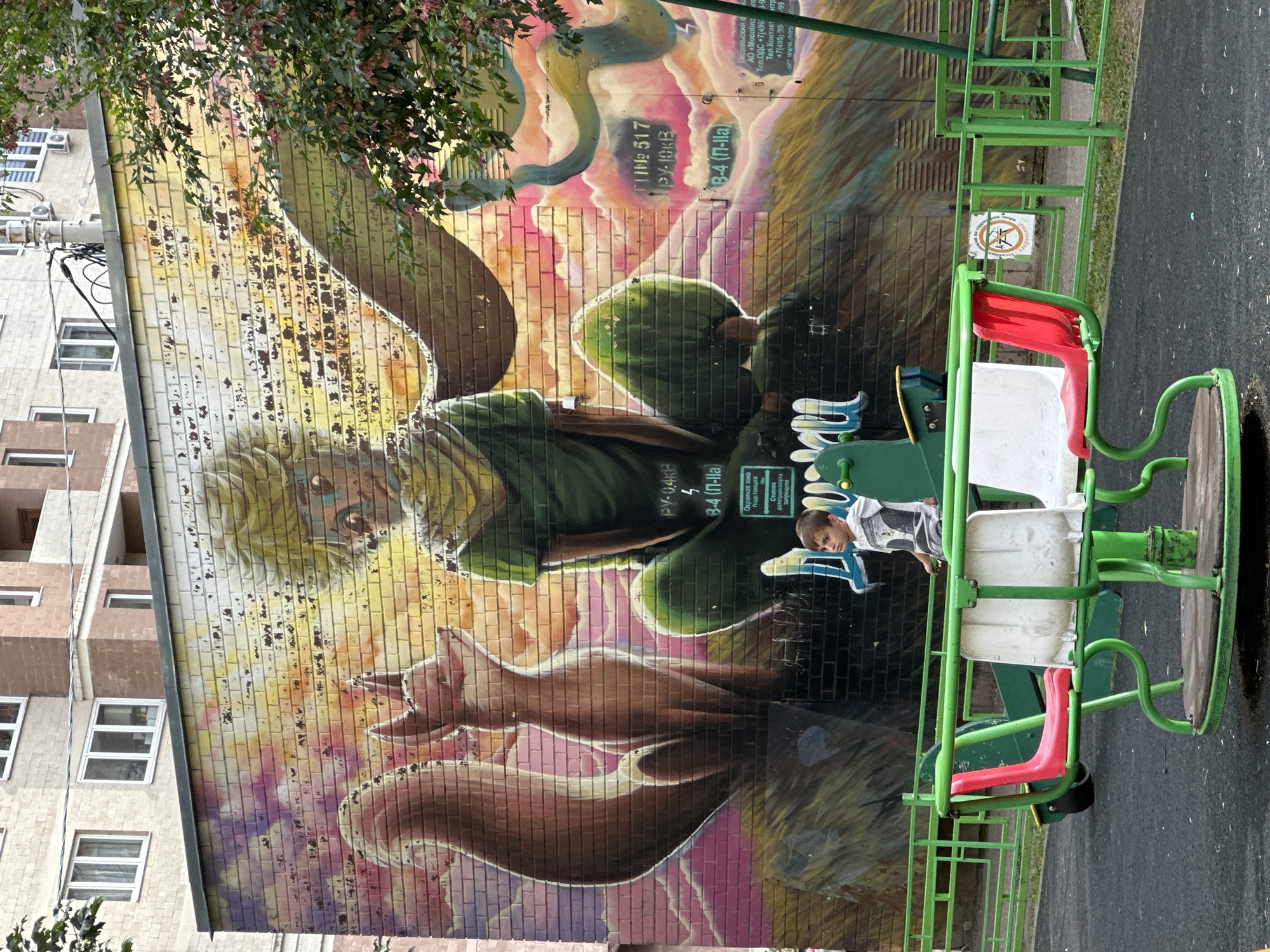 The image shows a vibrant mural on a brick building wall in Podolsk, Russia, depicting characters from "The Little Prince." The mural features the Little Prince with yellow hair and a green outfit, seated cross-legged, with a fox beside him. The background is a colorful depiction of clouds and a sunset. In the foreground, a child in a grey and white t-shirt sits on a green and red merry-go-round on a playground. The playground has a green metal fence and a black rubberized surface. The building behind the mural appears to be an apartment complex. There are some signs on the wall of the building near the mural.