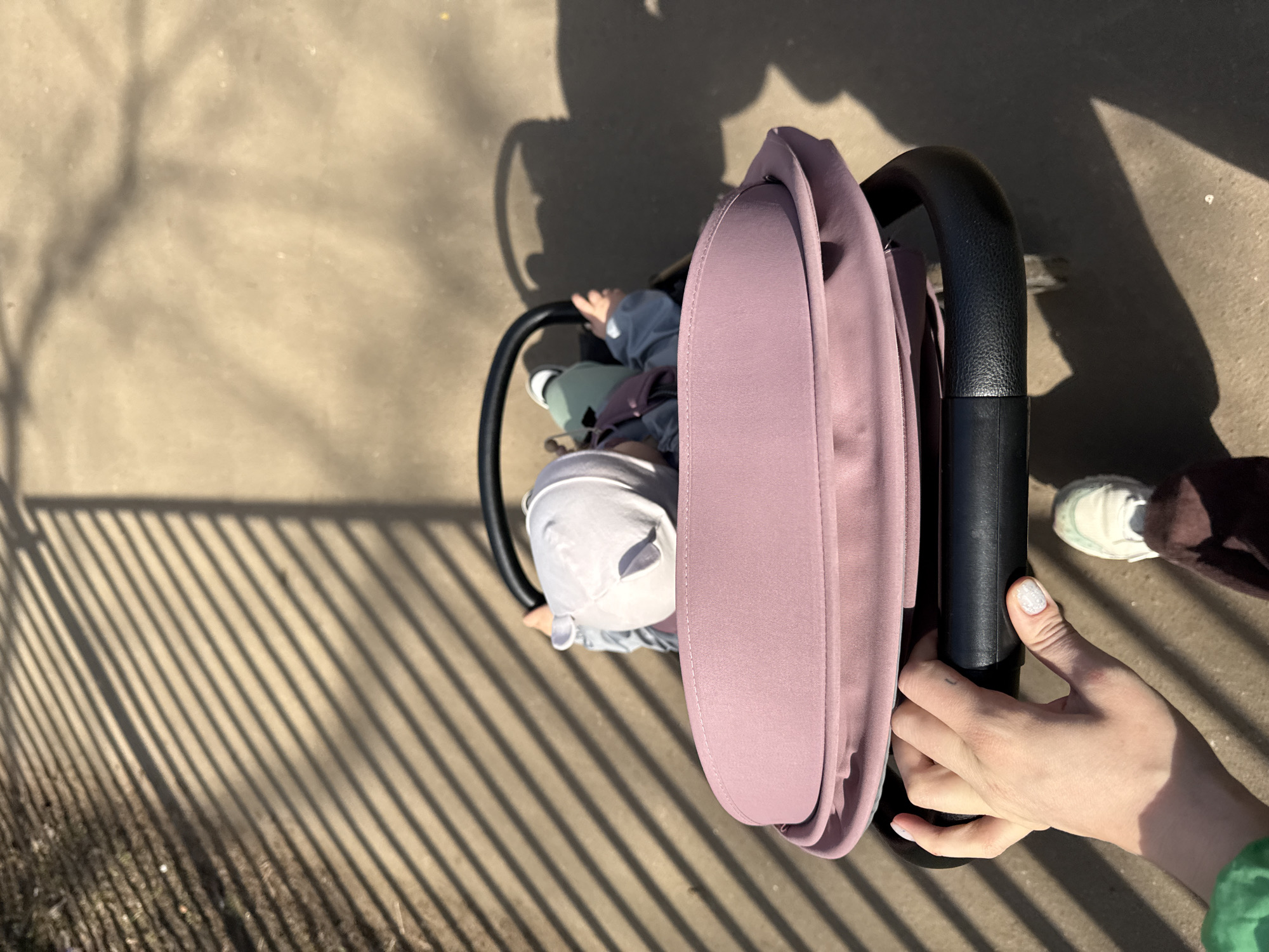 A top-down shot shows a baby in a stroller on a paved surface with strong shadows cast by a fence or barrier. The baby is wearing a light-colored hat and is partially visible inside the stroller. A person's hand is visible holding the stroller handle, with a shoe nearby. The scene is bathed in sunlight, creating high contrast shadows. The environment seems to be outdoors, potentially in a park or urban space. No information indicates the location is in Moscow, Russia.