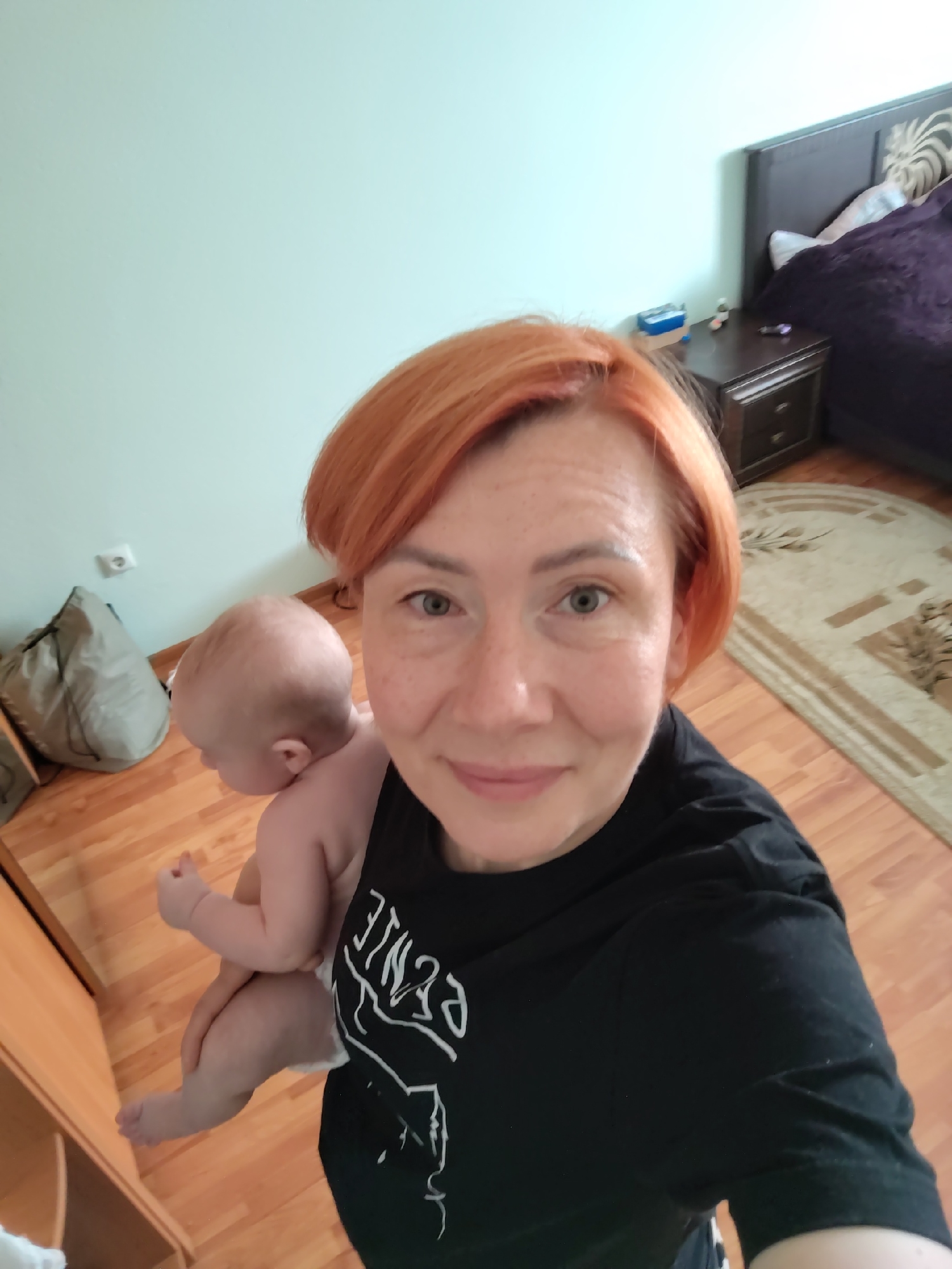 In Konakovo, Russia, a woman with short, reddish-orange hair and freckles smiles at the camera while holding a baby on her left shoulder. The baby is facing away from the camera, with its head tilted down. The woman is wearing a black t-shirt with white lettering. They are indoors, in a room with light blue walls and wooden flooring. To the left, a large, light gray duffel bag rests on the floor. In the background, a dark wooden bedside table with drawers is visible next to a bed with a dark purple duvet and patterned headboard. A rug with a geometric pattern is on the floor. The overall scene is a casual, indoor snapshot.