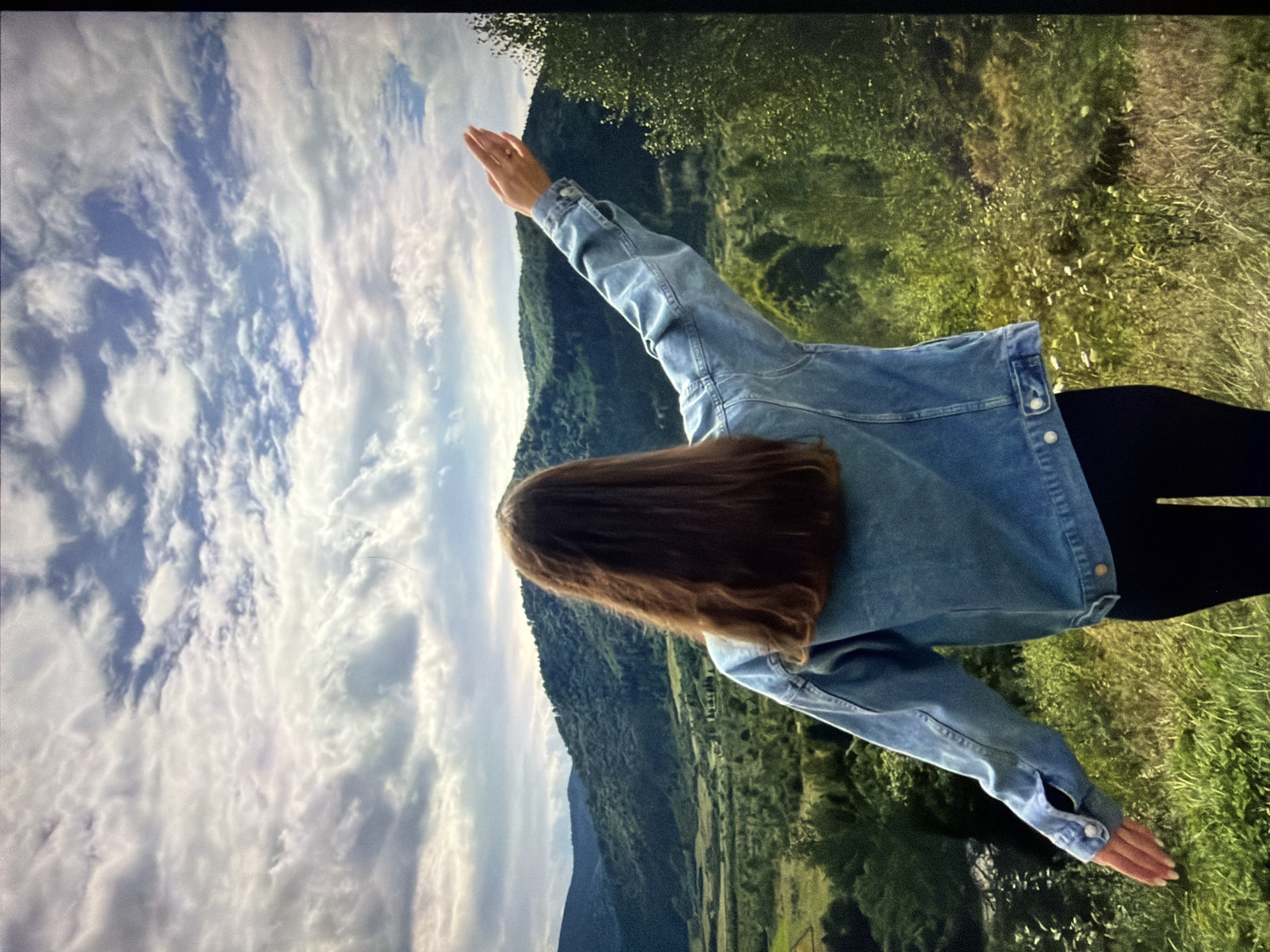 A woman with long, brown hair stands with her back to the camera, facing a landscape of rolling green hills and a dramatic sky filled with clouds. She is wearing a blue denim jacket and black pants, and her arms are outstretched to the sides, as if embracing the view. The environment is lush and natural, with trees and vegetation covering the hills. The sky is a mixture of blue and white, with sunlight breaking through the clouds in some areas. The scene evokes a sense of freedom and connection with nature. The image is a photograph taken outdoors.