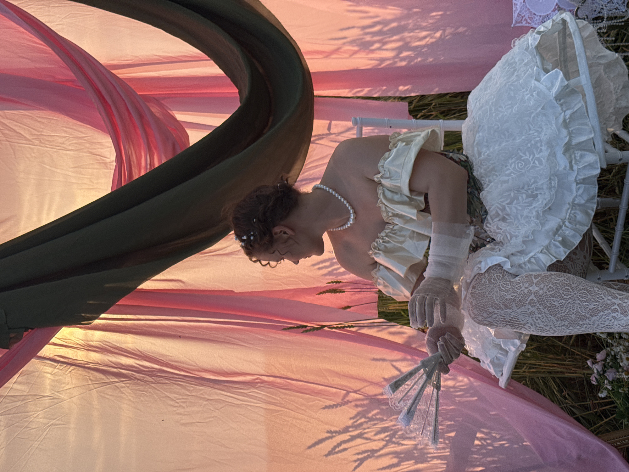 A person, likely female, is depicted in a stylized, almost theatrical pose within an outdoor setting. The subject is wearing a vintage-inspired outfit consisting of a pale yellow, off-the-shoulder top with ruffles, white lace stockings, and sheer white gloves. A pearl necklace adorns their neck. The subject holds a delicate, open fan made of white lace and pale colored metal.
The scene is characterized by flowing fabrics in shades of pink and a dark green. These fabrics are draped and bunched, creating a soft, ethereal backdrop that is illuminated by warm light, suggesting either sunrise or sunset. Shadows of what appear to be wheat stalks are cast across the pink fabric, indicating a rural or field location. Some small wildflowers are visible in the lower right corner. The subject is seated on a white chair, which is partially visible.
The overall mood is romantic and somewhat melancholic, enhanced by the soft lighting and the subject's contemplative posture, with their head bowed. The image conveys a sense of stillness and artistry, possibly a staged portrait or a scene from a narrative. The time of day is likely late afternoon or early morning, given the warm, low-angled light and pronounced shadows. The location context of Foki, Russia, suggests a rural or natural landscape. No text is visible in the image.