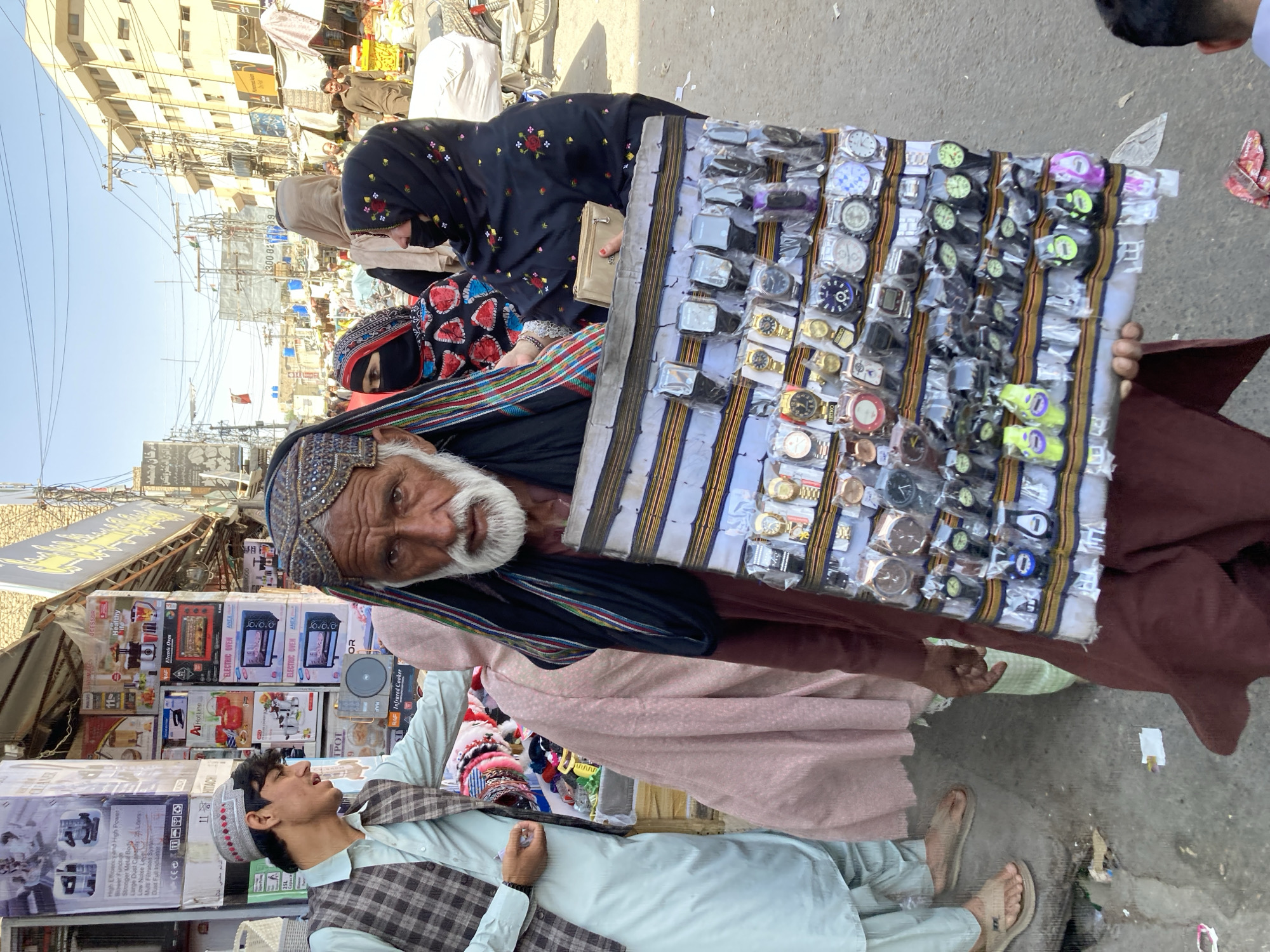 The image depicts a street scene in Quetta City Tehsil, Pakistan. An elderly man with a white beard, wearing a traditional head covering, holds a display of wristwatches. He faces the camera, engaging the viewer. Behind him, two women are seen, one wearing a black burqa with floral embroidery and the other also wearing a burqa. A young man in a light-colored shalwar kameez with a checkered waistcoat stands to the left of the elderly man. Behind him are stacks of boxes containing various appliances, including ovens and cookers. In the background, there is a crowded street with buildings, power lines, and various vendors. The overall environment appears to be a bustling marketplace.