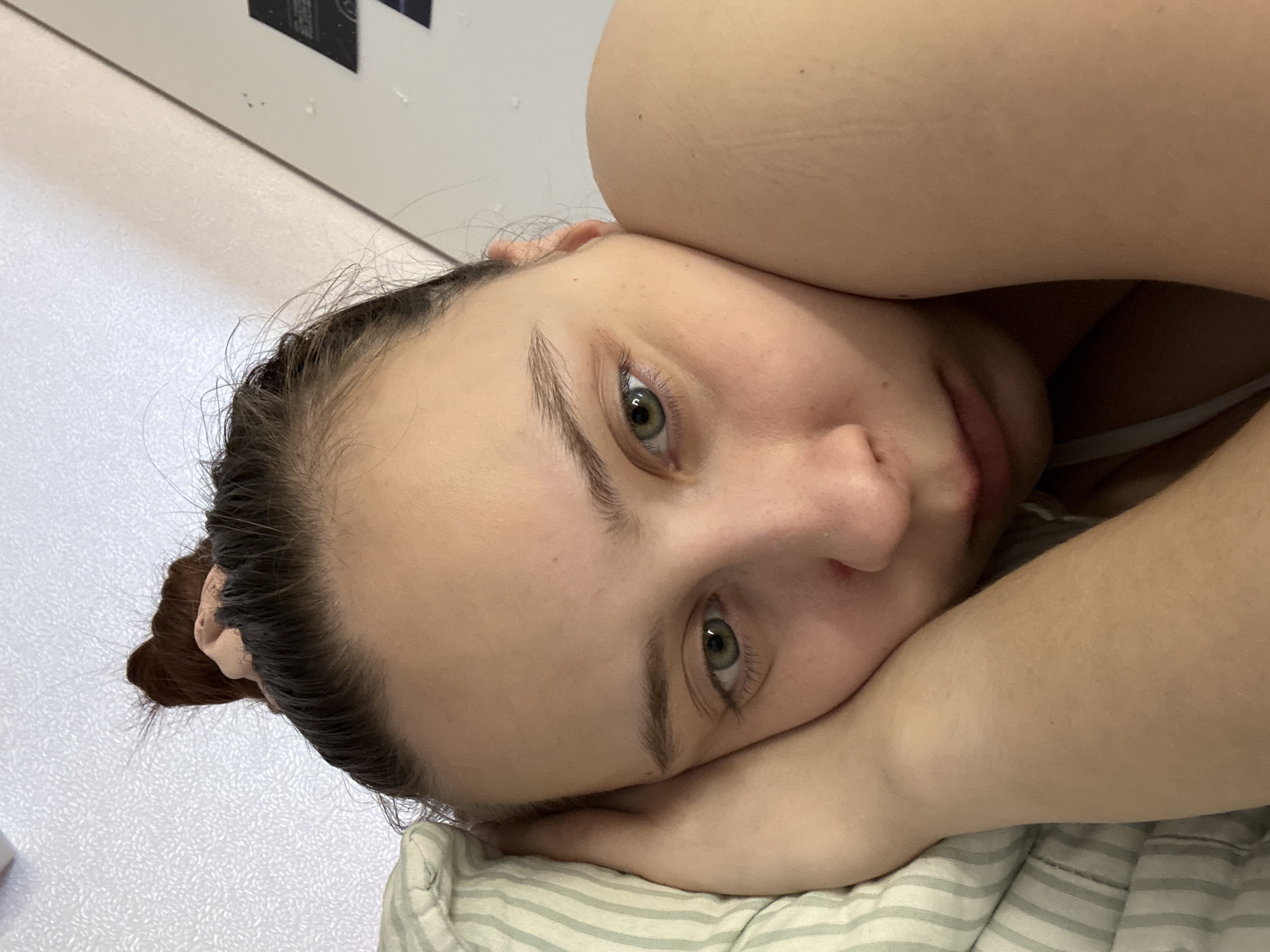 A selfie-style close-up photograph shows a young woman with fair skin, light green eyes, and dark hair pulled back into a bun. She is lying down, with her head resting on her folded arms. Her face is angled toward the camera. She is lying on a pillow with light green and white stripes. The background consists of a light-colored wall with an object, which could be an electrical fixture or switch, partially visible in the top left corner of the frame. There is no information to suggest the photo was taken in Прикамский, Russia.