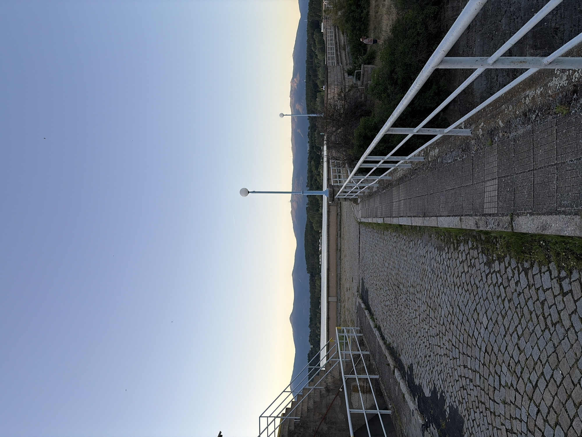 The image depicts a view of the Koprinka dam in Koprinka, Bulgaria, under a clear, pale blue sky. In the foreground, a cobblestone path leads diagonally towards the right, bordered by a white railing on the right side. To the left of the cobblestone path, there is a concrete structure with stairs and a white railing. Beyond the immediate foreground, a paved walkway with a similar white railing stretches forward. In the middle ground, there is a body of water, and across the water, a densely forested area is visible, leading to a mountain range in the distance. Two lampposts with spherical lights stand on the paved walkway. A person is standing on a grassy area to the right of the paved walkway, in the middle distance. The overall scene suggests an outdoor public space, likely a promenade or viewing area associated with the dam.