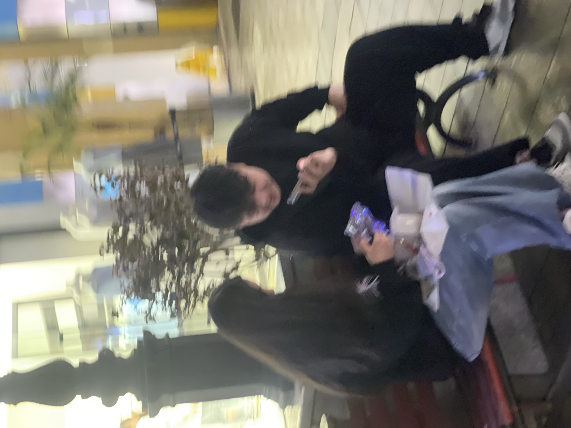 The scene depicts two individuals seated on a red-brown slatted bench in an urban environment, identified as the Chongqing High-tech Zone, China. The setting appears to be an outdoor or semi-outdoor pedestrian area, illuminated by artificial light, possibly at night.
On the left, a female individual with long, dark hair wears a dark top with a light-colored design and light blue wide-leg jeans. She is focused downwards, holding a clear object with internal blue illumination and a white, possibly packaged, item.
On the right, a male individual with dark, short hair wears a dark long-sleeved top and dark pants, paired with light-colored sneakers. He is holding a smartphone in his left hand, looking at its screen.
In the blurred background, a dark, ornate pillar is visible to the far left. A small tree or bush with dark foliage stands between the pillar and the seated individuals. Further back, brightly lit shop windows display mannequins or abstract forms. A yellow "wet floor" or caution sign is faintly discernible in the far right background. The ground beneath the bench appears to be a paved surface. Both individuals are engaged with their respective handheld devices, without apparent direct interaction with each other.
