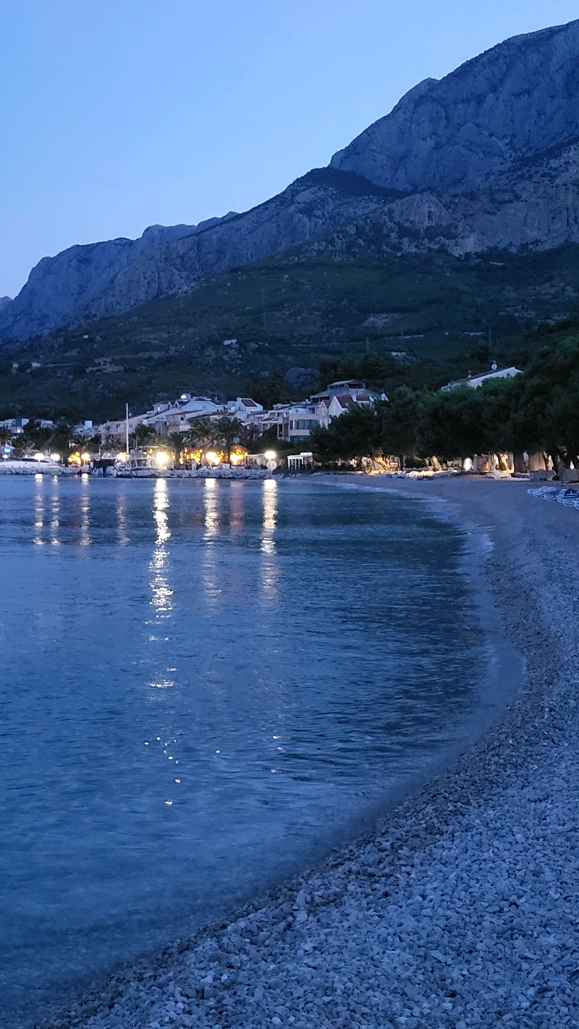 The image shows a twilight scene at a pebble beach with calm, dark blue water reflecting the lights of a nearby town. The town consists of buildings with tiled roofs and illuminated windows and streetlights, situated along a waterfront. Palm trees and other vegetation are present between the town and the beach. In the background, a large, dark mountain range with visible rocky textures rises steeply. The sky is a clear, deep blue, typical of dusk. The beach itself is covered in small, light-colored pebbles. No people are visible in the image. The overall atmosphere is serene and peaceful. This location is likely along the coast of Općina Tučepi, Croatia, given the characteristic Adriatic landscape of mountains meeting the sea and the architectural style of the buildings.