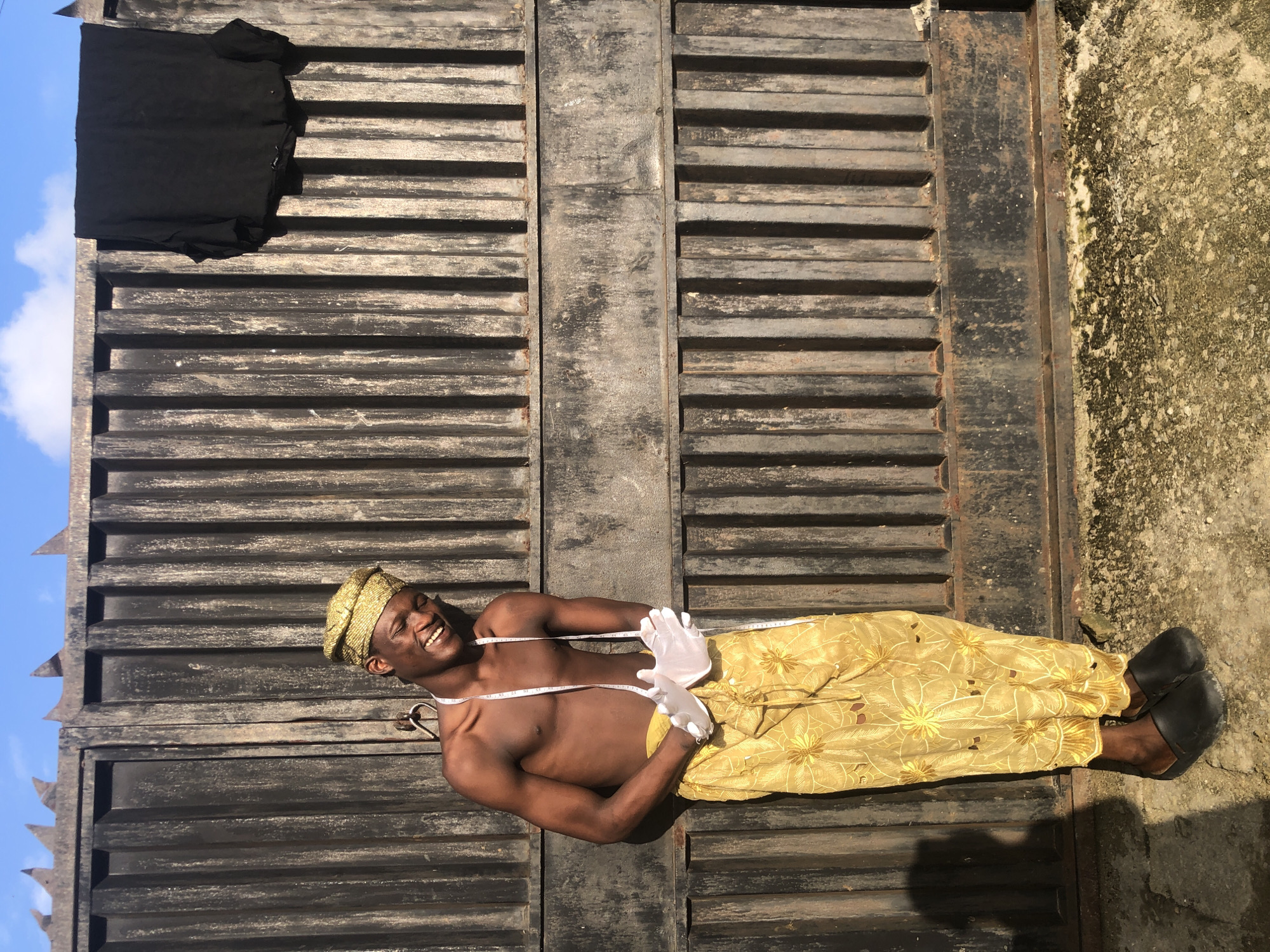 A young Black male stands outdoors in front of a dark, corrugated metal gate in Ado, Nigeria. He is shirtless and wears a gold-colored, textured headwrap and a golden, patterned cloth wrapped around his lower body. White gloves cover his hands, which are clasped together in front of him. A white tape measure is draped around his neck and extends down his chest. The gate is weathered and has pointed spikes along its top edge. A black cloth is draped over the top of the gate on the right side. The ground in front of the gate is a mixture of concrete and dirt, with some moss and debris. The sky above is blue with scattered white clouds.