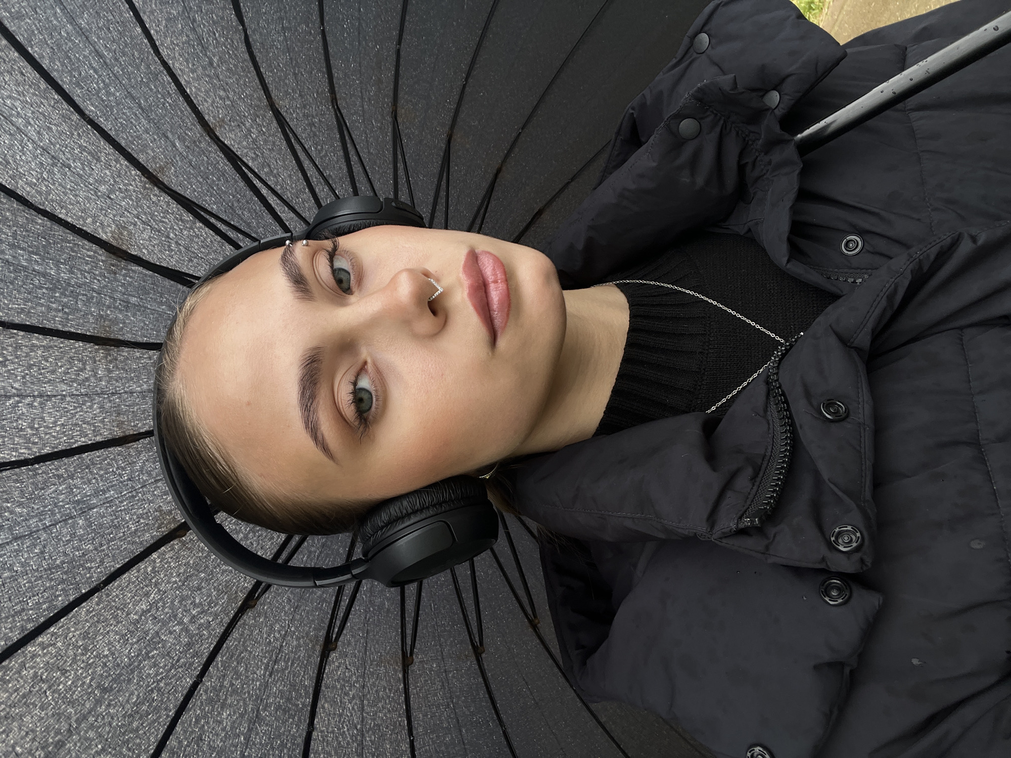 A young woman is depicted lying down, looking directly at the camera with a neutral expression. She is wearing black over-ear headphones, a black puffer jacket, and a black knitted sweater, along with a silver chain necklace. Her light-colored eyes (greenish-gray) and dark eyebrows are prominent. She has an eyebrow piercing with silver beads and a rhinestone-adorned hoop piercing in her nose. Her lips have a subtle pink tint.
The woman is positioned on or in front of a large, opened black umbrella, which serves as a textured, dark background, displaying its ribs and canopy material. The umbrella appears to be wet or have water droplets on it, and similar droplets are visible on her puffer jacket in some areas, suggesting recent or ongoing rain. In the upper right corner of the original image, a glimpse of an outdoor setting, possibly pavement or ground, can be seen, indicating the photo was taken outdoors during the daytime, likely under overcast conditions.
The scene takes place in Фоки, Russia. No visible text is present in the image.