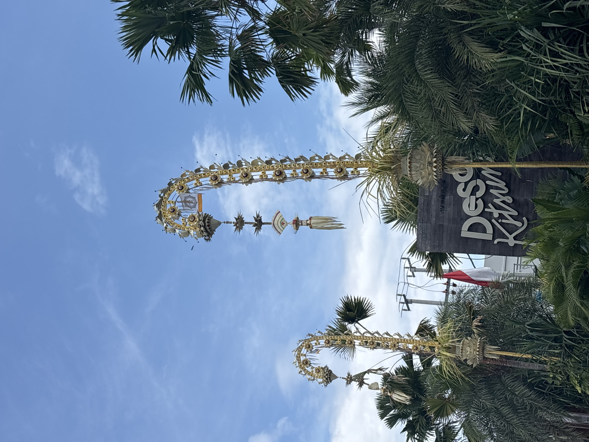 The image is taken in North Kuta, Indonesia and is oriented to look upwards at two ornate, curved decorations that appear to be made of gold or yellow material, along with white fringe, each positioned atop a tall palm tree. The background is a bright blue sky with sparse clouds. To the right of the top decoration, there is a wooden sign that says "Desa Kitsin". Adjacent to the sign are palm trees and what looks like a red and white flag. In the background, utility poles and power lines can be observed.