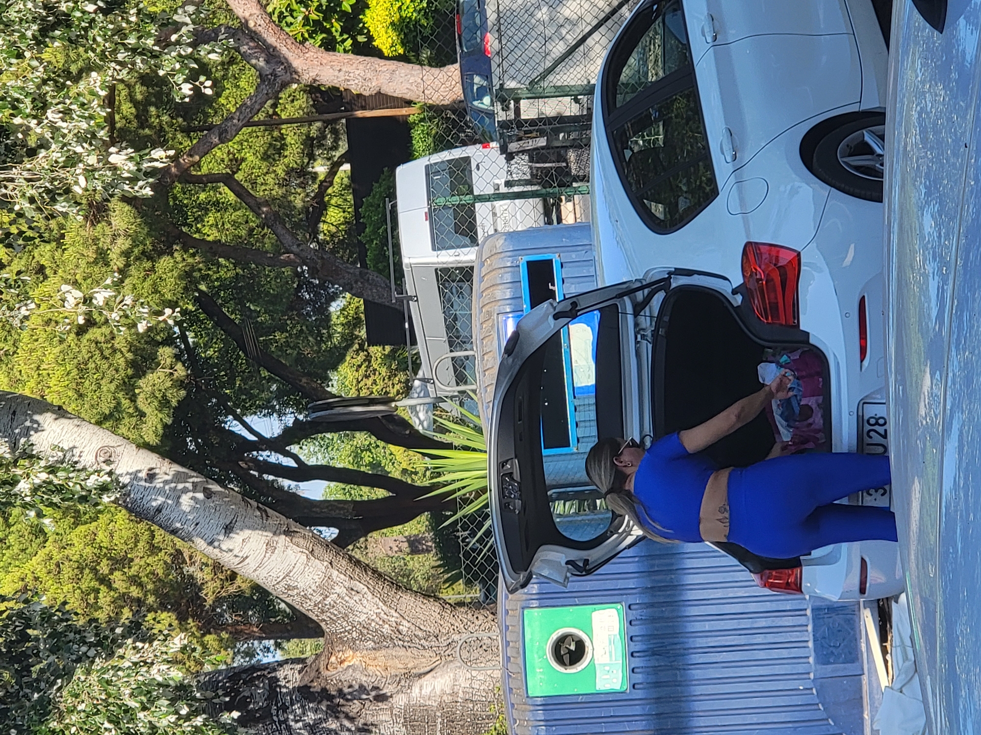 A woman in a blue athletic outfit with her hair tied back in a ponytail is standing at the open trunk of a white sedan, retrieving items from a colorful bag. She has a tattoo on her lower back. The car is parked next to a large blue recycling bin with a green circular aperture for glass disposal. In the background, a white van is visible, along with other vehicles behind a chain-link fence. Lush green trees and foliage surround the scene, suggesting an outdoor setting. The license plate of the white sedan is partially obscured, showing the characters "J28". The environment appears to be a public area in Quarteira, Portugal, likely a recycling point.