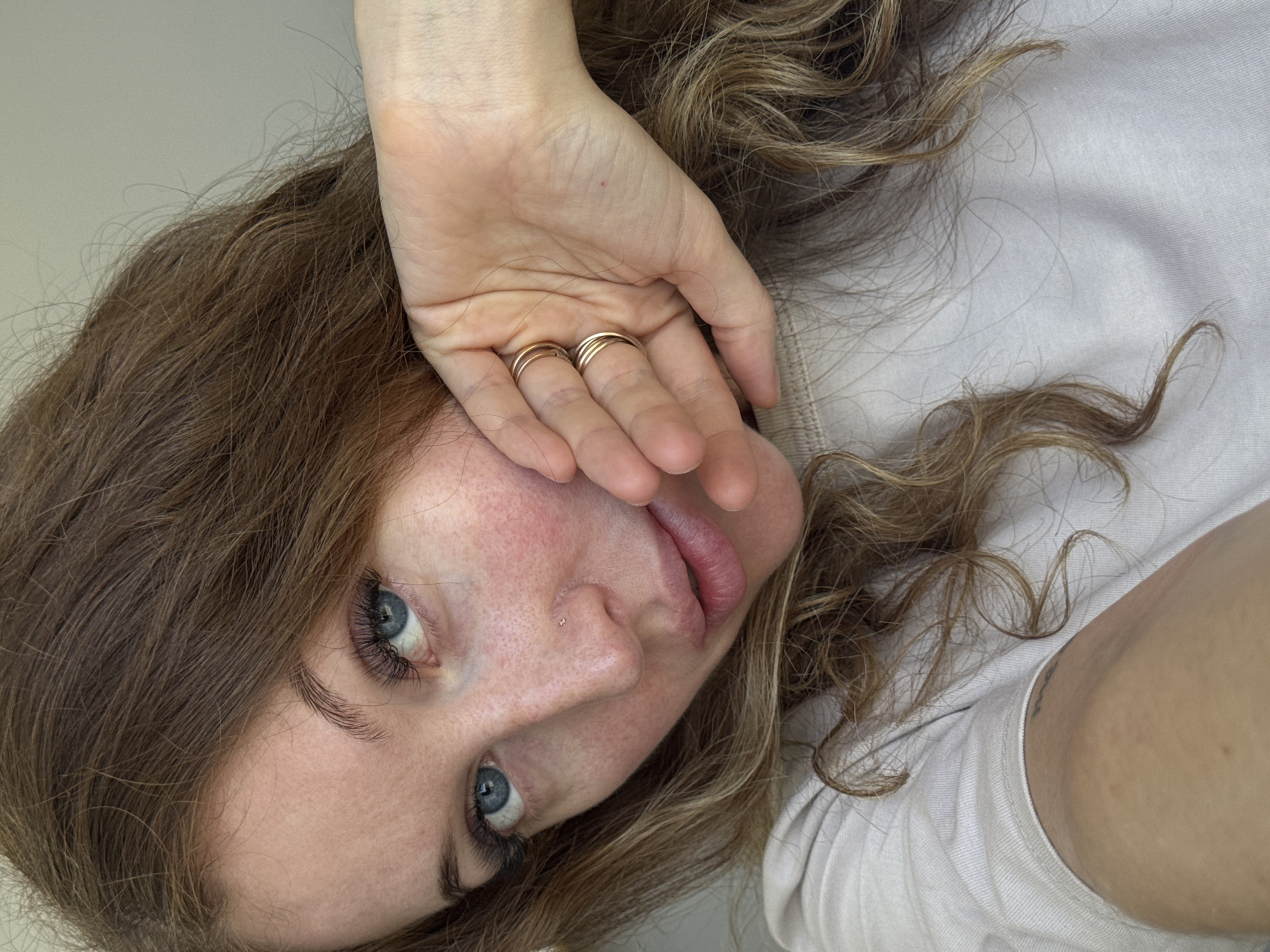 A woman is lying on her side with her head tilted towards the camera. Her face is partially obscured by her hand, which has two gold rings on two of the fingers. She has fair skin, light brown hair with blonde highlights, and blue eyes. A small piercing is visible on her left nostril. She is wearing a light-colored t-shirt. A tattoo is visible on the arm. The background is a plain, light-colored surface. No details confirm the location in Chaikovsky, Russia.