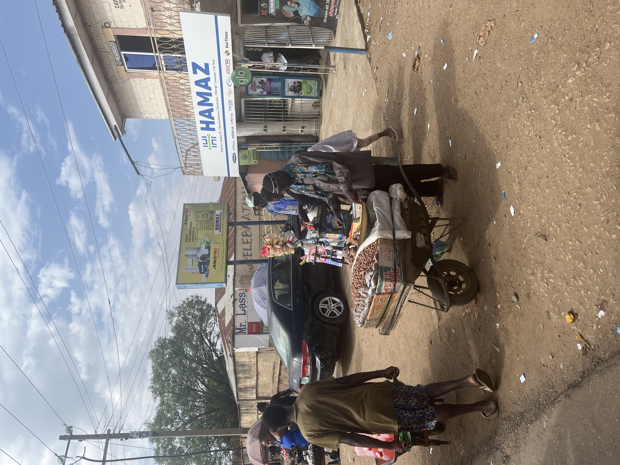 The image shows a bustling street scene in Jos, Nigeria. A vendor with a cart laden with assorted items, including bags of peanuts and various goods, stands next to a parked black car. Two men appear to be examining or engaging with the vendor's wares. The street is littered with small pieces of paper or debris. Buildings and signs bearing the name "HAMAZ" are visible in the background, advertising telecommunications, electronics, and related services. Additional signs advertise Mr. Lass, Coca-Cola, Mewe appliances, Glo, and others. Trees and power lines are also present.