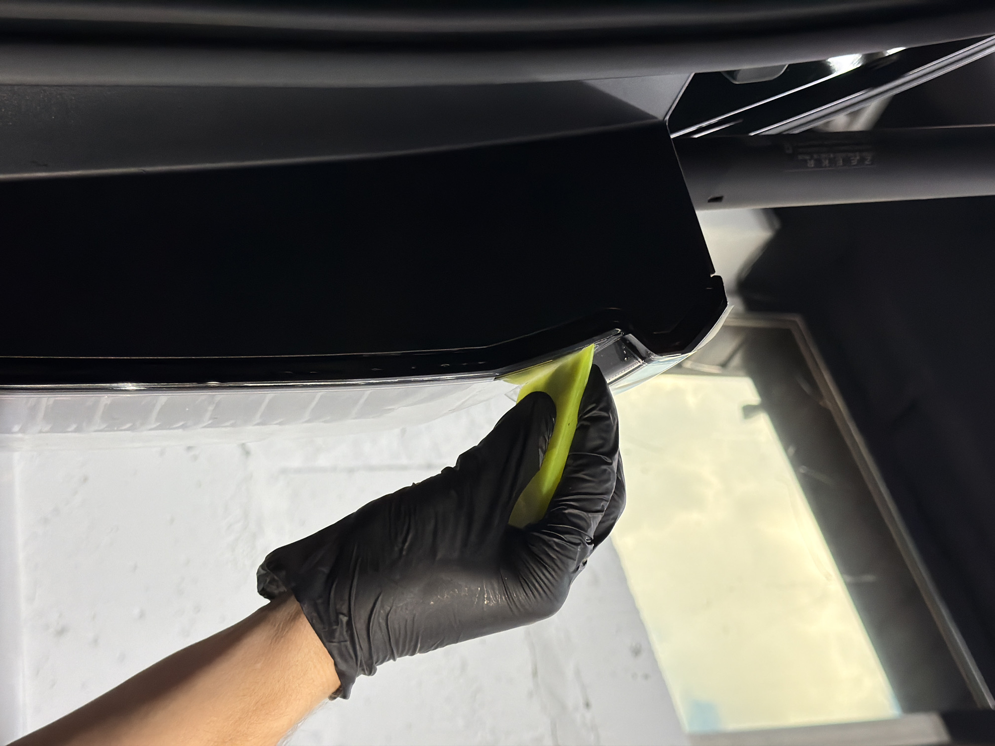 A close-up, low-angle shot shows a gloved hand with a yellow squeegee pressing against the edge of a black car's exterior trim. The gloved hand is wearing a black nitrile glove. The background consists of a window reflecting the sky, and a white, textured wall. The image is taken indoors, likely in a car detailing or repair shop. There is no indication of the location being Krasnoyarsk, Russia.