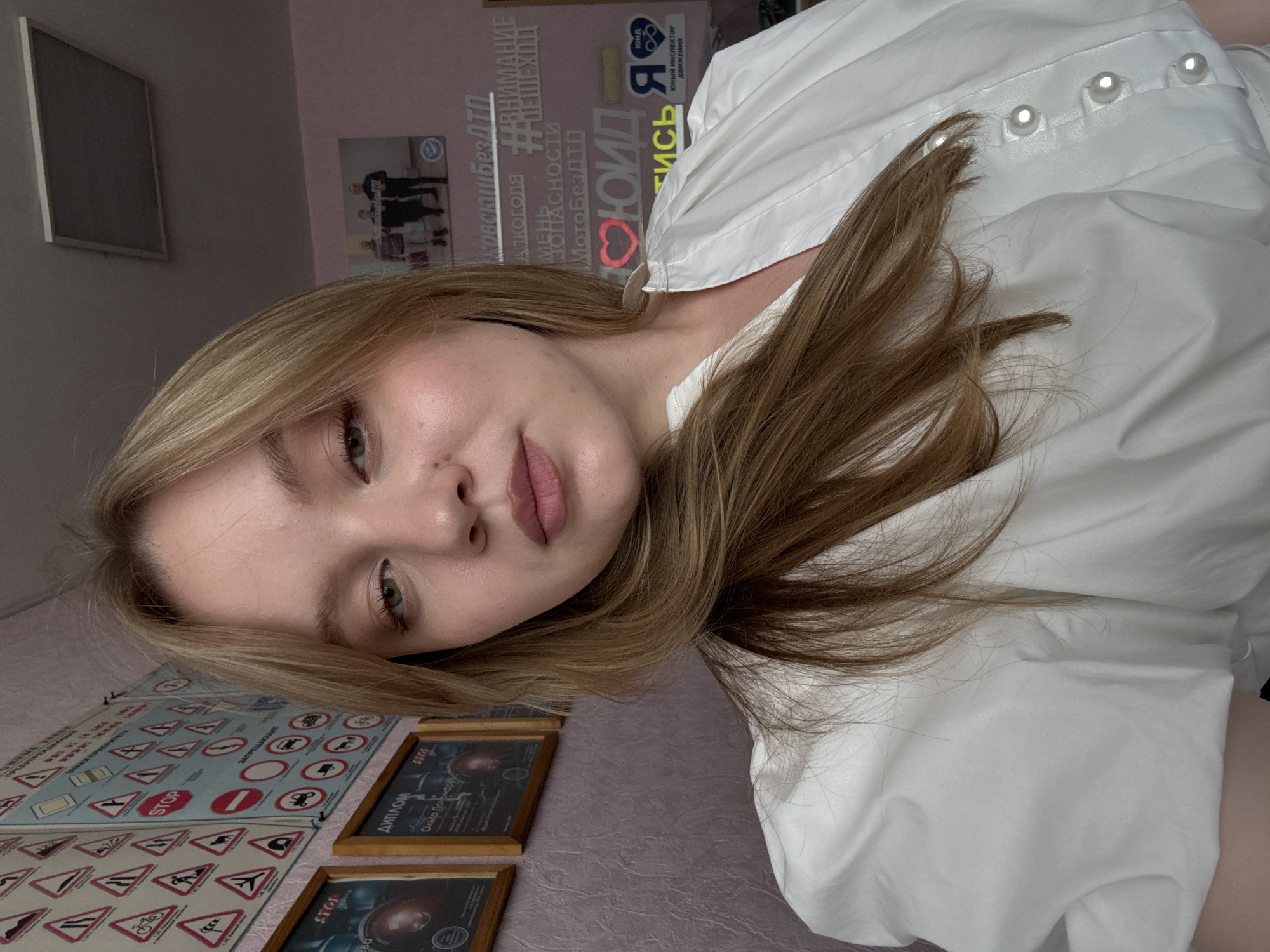 A young woman with blonde hair, wearing a white blouse with pearl buttons, poses for a selfie in a room with pink walls. The wall behind her is adorned with various road signs and framed certificates, suggesting the location might be a driving school or a similar educational institution. Text visible on the wall includes "STOP," "ДИПЛОМ" (Diploma), and Russian phrases related to traffic safety, such as "ВНИМАНИЕ" (Attention), "АЛКОГОЛЯ" (Alcohol), and "МОТОБЕЗОПАСНОСТИ" (Motorcycle Safety). A small framed photograph of three individuals is also visible on the wall. The lighting suggests an indoor setting. The presence of the Russian text and the specific road signs could indicate a location within Russia, potentially Chaikovsky, Russia, given the information.