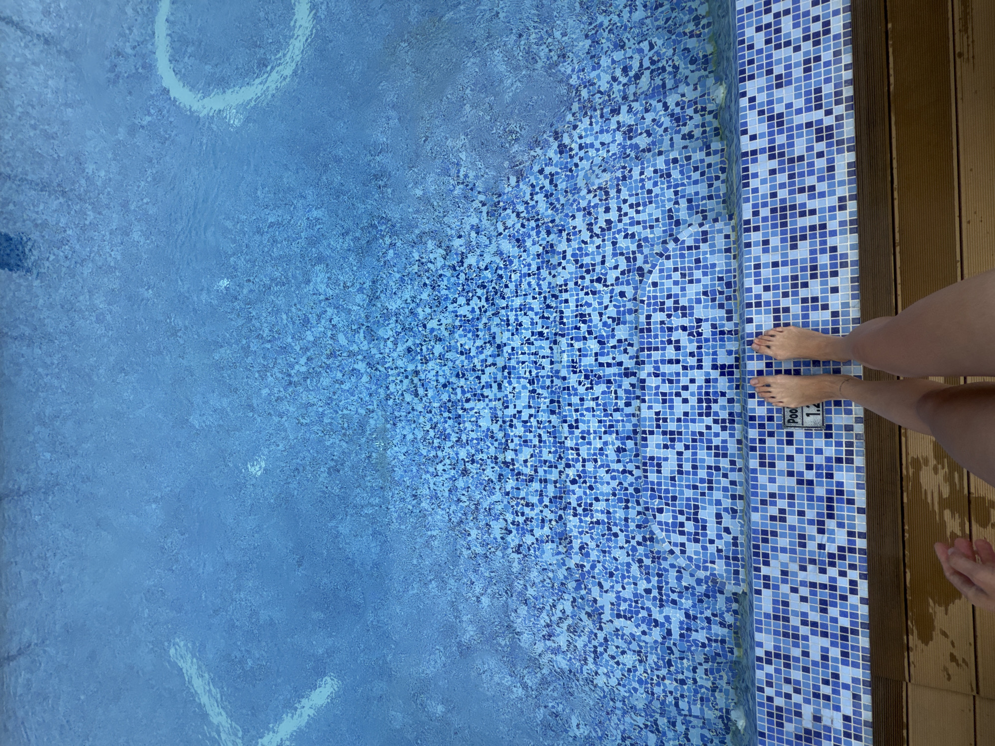 The image depicts an overhead view of the edge of a swimming pool in North Kuta, Indonesia. The pool features a tiled bottom composed of small blue and white mosaic tiles, with the water clarity allowing the tiles to be visible. In the foreground, a person's bare feet are visible, positioned at the edge of the pool, with the lower legs and thighs also in frame. A sign on the pool edge indicates "Pool 1.2", likely referring to a depth of 1.2 meters. The pool area transitions to a wooden deck. There is water on the wooden deck, indicating a wet environment. Large white lettering is visible submerged in the pool water, but the full text is not clear.