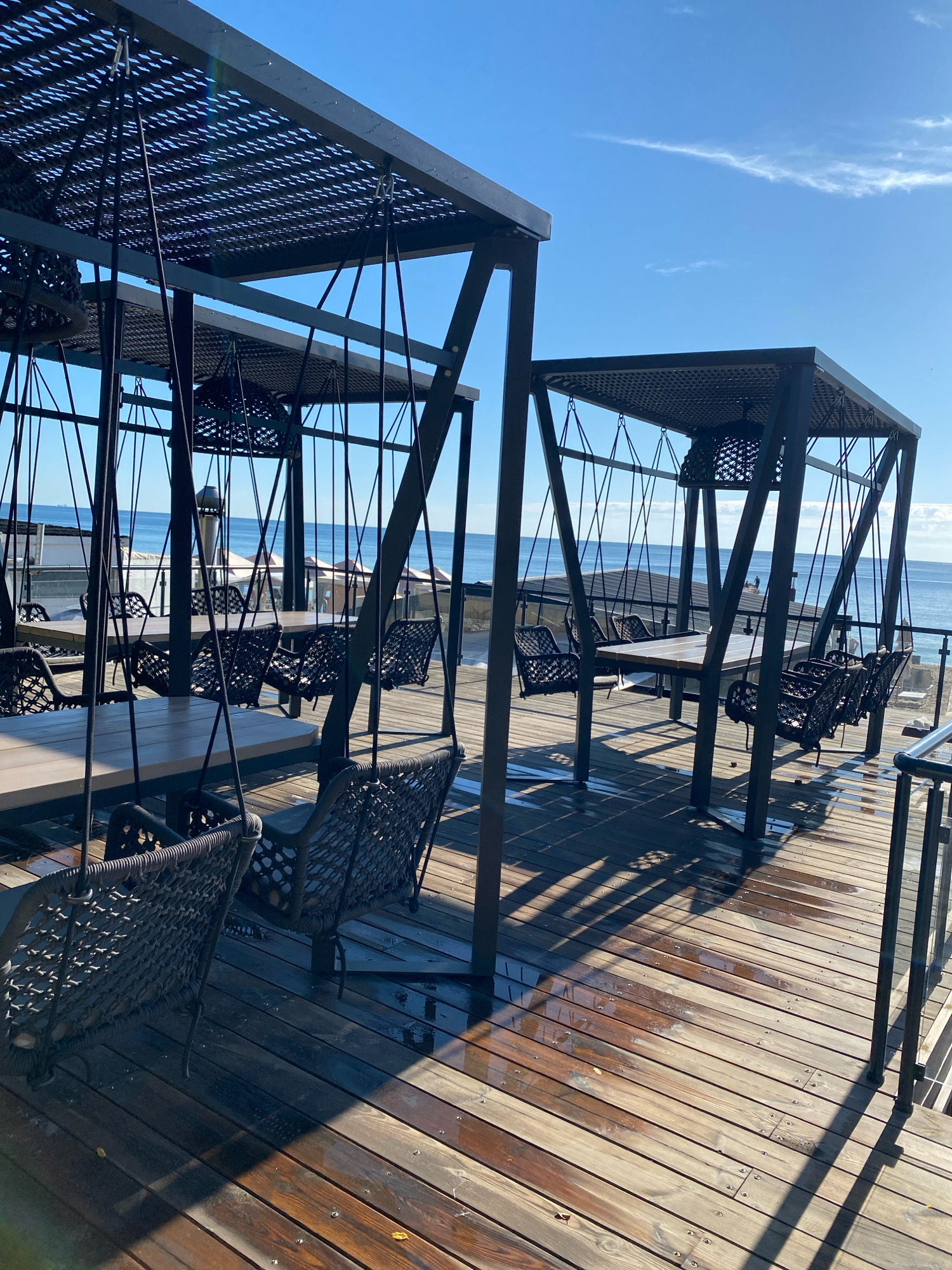 This is a photograph of an outdoor seating area near the sea in Odesa, Ukraine. The seating area features modern, dark-colored pergolas with perforated roofs, under which are tables and chairs. Some of the chairs are suspended as swings, crafted from woven material. The floor of the seating area is a wooden deck, which appears wet in places, reflecting the sunlight and shadows. Beyond the deck, the blue sea stretches to the horizon under a clear sky with a wispy cloud. In the distance, along the coastline, there are glimpses of buildings and other outdoor structures, possibly part of a resort or restaurant. The overall atmosphere is tranquil and suggests a place designed for relaxation by the sea.