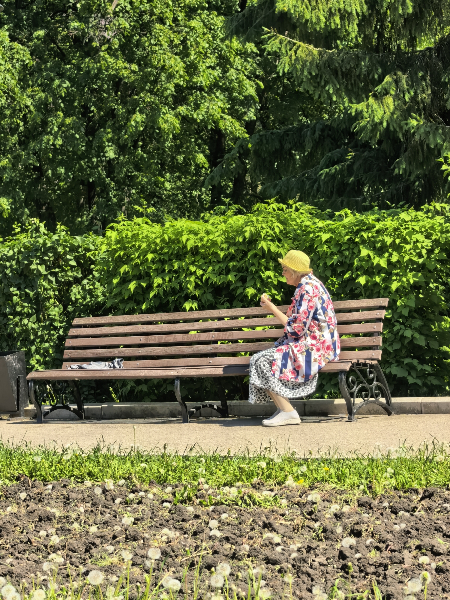 An elderly woman wearing a yellow hat, a floral shirt, and a patterned skirt sits on a wooden bench in a park. She appears to be holding and looking at something in her hands. The bench is located on a paved path. In the foreground, there's a patch of earth with sparse grass and dandelions. Behind the bench, there is a dense green hedge and trees. The location is Sterlitamak, Russia.