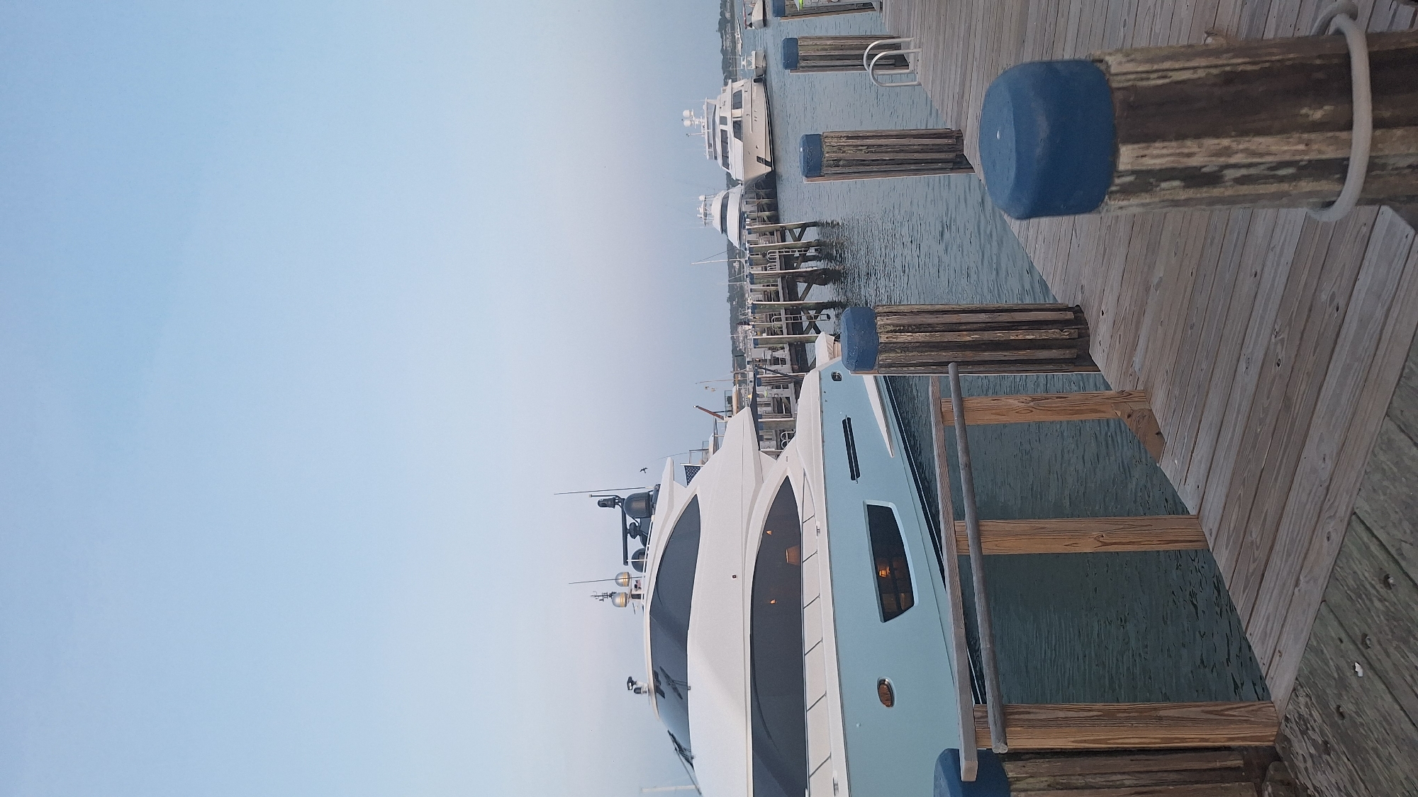 The image shows a view of boats docked at a harbor in Nantucket, USA. In the foreground, a wooden pier with vertical posts topped with blue caps leads into the water. A light blue and white yacht is prominently docked on the left side of the frame. In the background, several other yachts are docked along a pier. The sky is clear and pale blue, indicating it might be early morning or late evening. The water is calm and reflects the light from the boats and the sky. The overall scene depicts a serene harbor setting with luxury yachts.