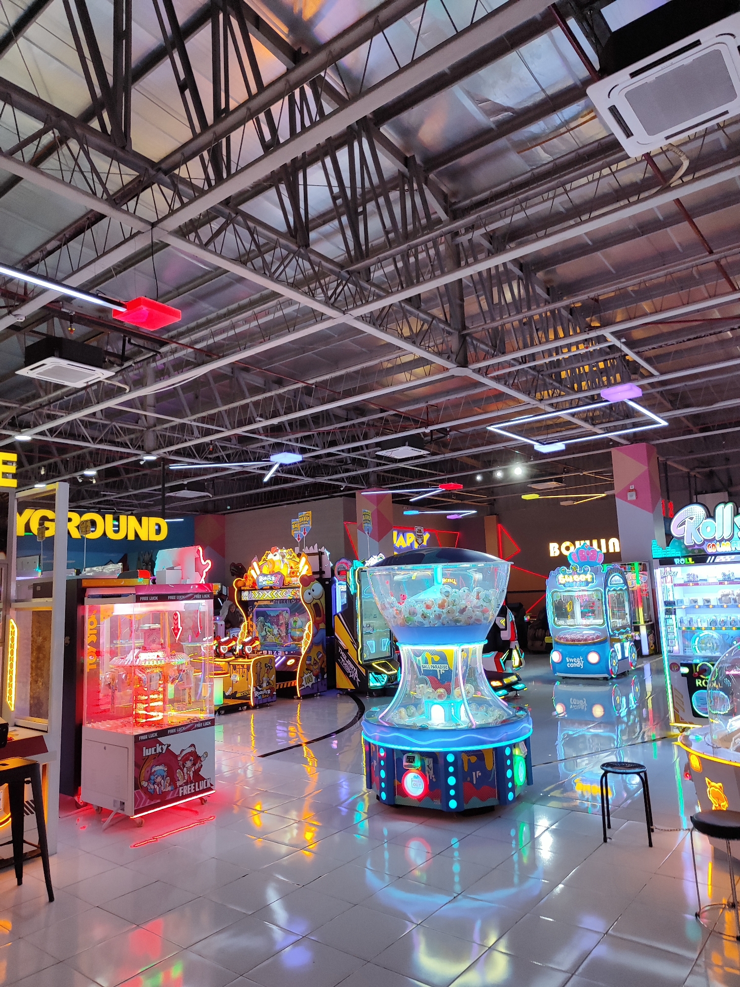 This image depicts the interior of an arcade in Sempusari, Indonesia. The space is brightly lit with a variety of neon and LED lights emanating from arcade machines and ceiling fixtures. The ceiling is high and features exposed industrial beams and ductwork. The floor is tiled and reflects the colorful lights.

Several arcade machines are visible, including claw machines, redemption games, and video games. One prominent machine in the foreground is a claw machine with a red neon base and "Lucky Free Luck" branding. To its right, a large, cylindrical machine with a clear top filled with small balls and illuminated with blue LEDs, labeled "BALL PARADISE," is present. Further into the background, more machines are visible, including one with a "Sweet candy" label and another that says "Roll." Signs with text such as "PLAYGROUND" and "BOHLIN" are also visible on the walls.

There are no people clearly visible in the image, though the presence of the arcade suggests it is a place for entertainment. The overall impression is a lively and modern entertainment venue.