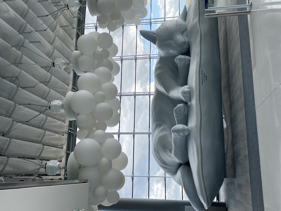 This image captures a large, light-gray sculpture of a sleeping cat on a matching pillow, situated within Terminal 2E of Charles de Gaulle Airport, near Mitry-Mory, France. The oversized cat sculpture is depicted in a relaxed, curled pose with its eyes closed. Text inscribed on the front of the pillow reads "DÉFENSE DE MONTER," "NO CLIMBING," and Japanese characters. The sculpture rests on a low, rectangular base of the same light gray color.
The environment features a high, vaulted ceiling with a white, ribbed or paneled surface. Suspended from the ceiling are numerous clusters of white, spherical light fixtures varying in size, resembling abstract clouds. A prominent multi-paneled glass window wall forms the background, revealing a bright, overcast sky with visible clouds. The floor appears to be a large-tiled or carpeted surface, primarily gray with lighter gray accents. A dark structural column is partially visible on the far left, and a silver stanchion barrier is present on the far right, indicating a public interior space. No people are visible.