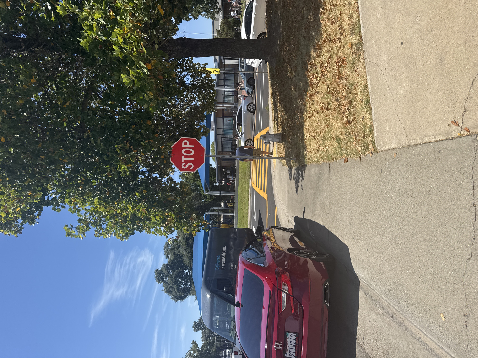 A red Honda Accord is parked on a sidewalk, with its rear facing the camera. To the left of the Accord, a black Amazon Prime delivery van is parked. In the background, a street intersection with a stop sign is visible, along with several cars and people. The scene appears to be in front of a school, with a fenced playground area and a building in the background. The sky is clear and blue, and trees with green leaves are present. The ground is paved with concrete sidewalks and asphalt roads, with patches of dry grass and fallen leaves. The location is identified as Citrus Heights, USA.