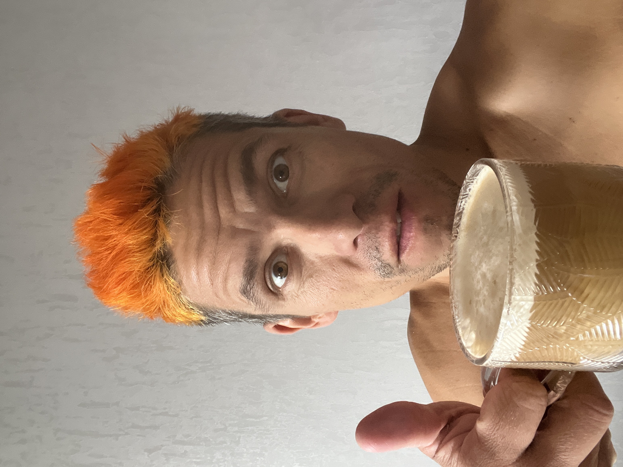 A person with bright orange hair is shown from the chest up, holding a glass mug of coffee with a frothy top. The person's thumb is extended towards the camera. They are looking directly at the camera with wide eyes and their mouth slightly open. The background is a plain, textured light gray wall. The image appears to be a selfie taken indoors. There is no discernible information in the image that would indicate its location in the city of Chaikovsky, Russia.