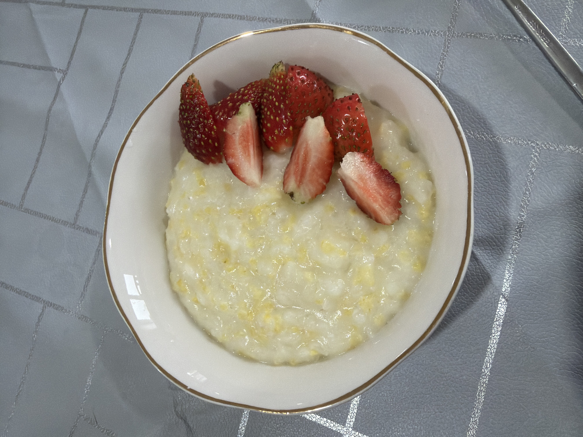 A white bowl with a gold rim sits on a light gray textured tablecloth. Inside the bowl is a creamy, pale yellow porridge. Several sliced and whole strawberries are arranged on top of the porridge. The strawberries are bright red with visible seeds. The tablecloth has a subtle diamond pattern with thin, reflective silver lines. A portion of a silver utensil, possibly a spoon, is visible in the lower right corner, with the words "STAINLESS STEEL" embossed on it. The image is taken from a top-down perspective. There are no people or specific landmarks of Chaikovsky, Russia visible in the image.