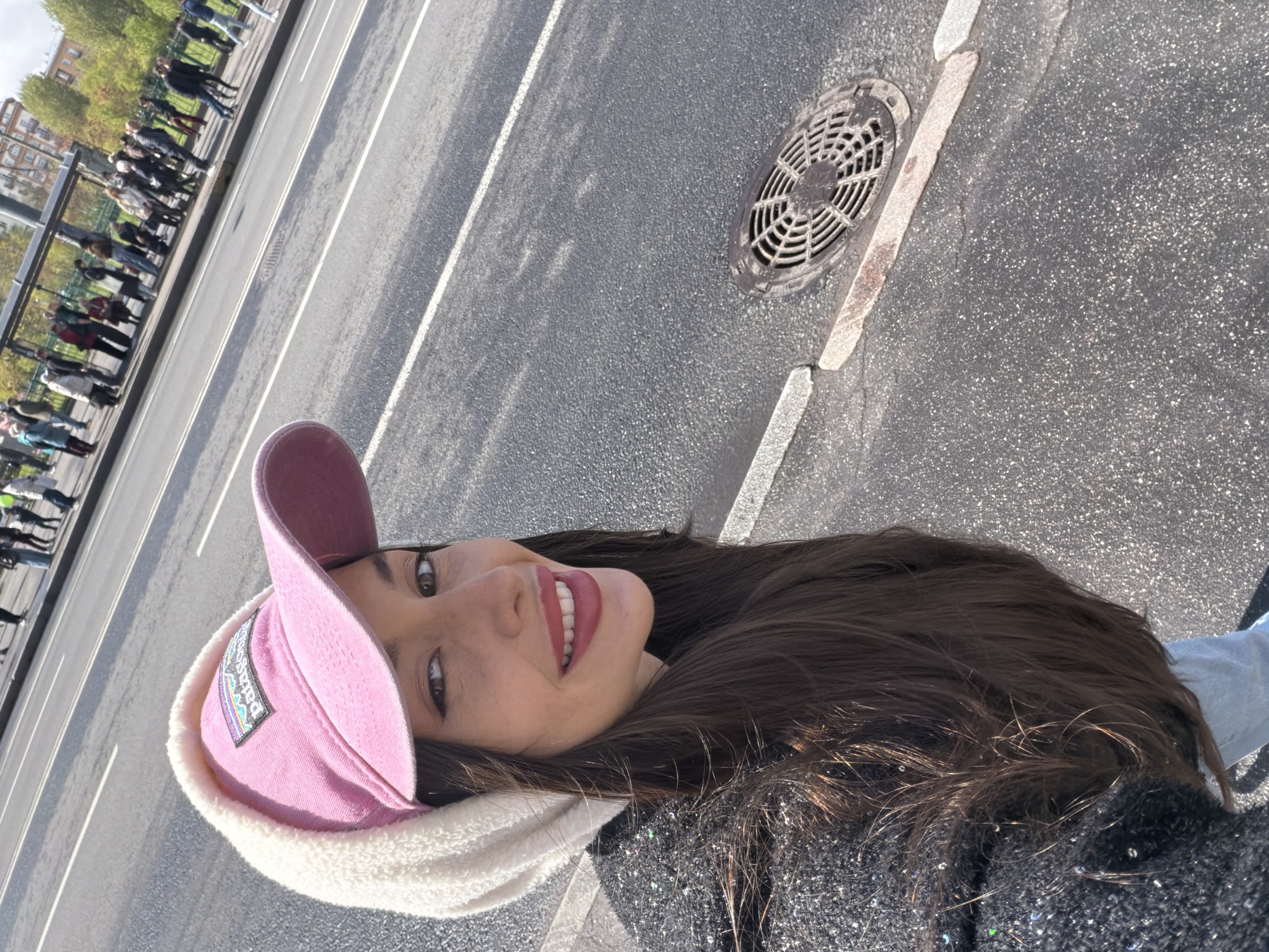 A young woman wearing a pink baseball cap with a white sherpa brim smiles at the camera. She is standing on an asphalt sidewalk next to a road with a manhole cover. Across the road is a bus stop with a line of people waiting. This location is in Saint Petersburg, Russia.