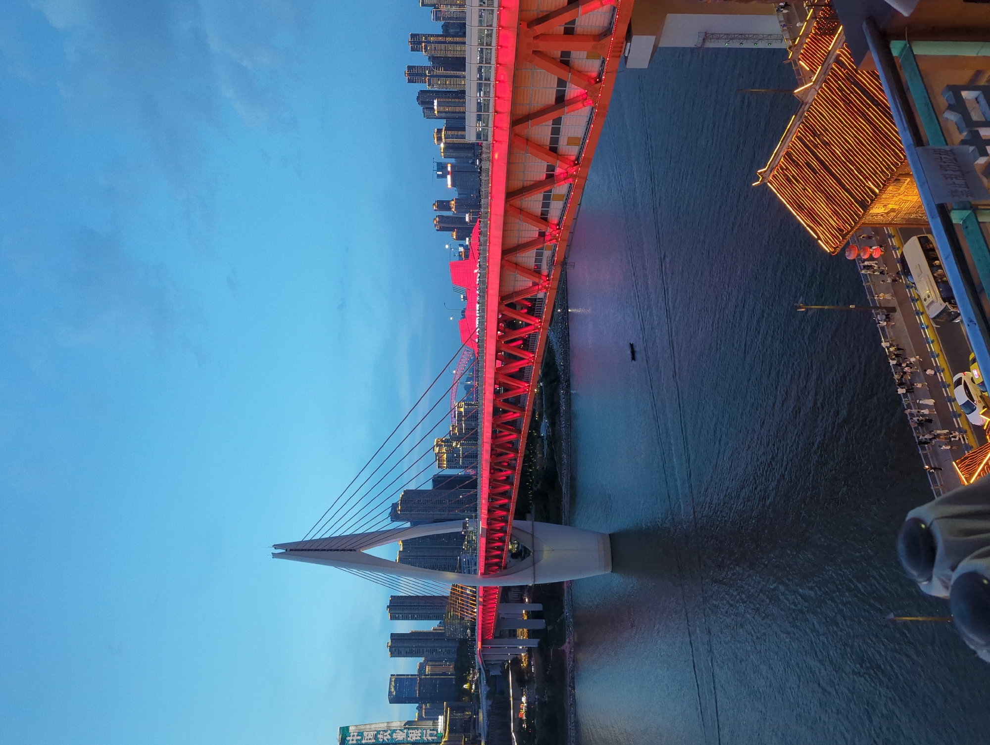 The image shows the Hongyadong Bridge in Yuzhong District, China, at dusk. The bridge, illuminated in bright red, spans a wide body of water. The cable-stayed design features a prominent central pylon. In the background, a cityscape of modern skyscrapers with illuminated windows rises against the twilight sky. On the waterfront, traditional Chinese architecture with illuminated roofs is visible, as well as a pedestrian walkway with people strolling and a white bus. The water reflects the lights from the bridge and the city. A small boat is present on the water.